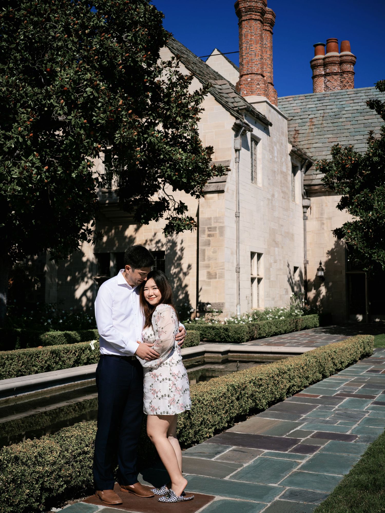 A couple embraces on a stone pathway outside a Gothic-style building with tall chimneys and manicured hedges.