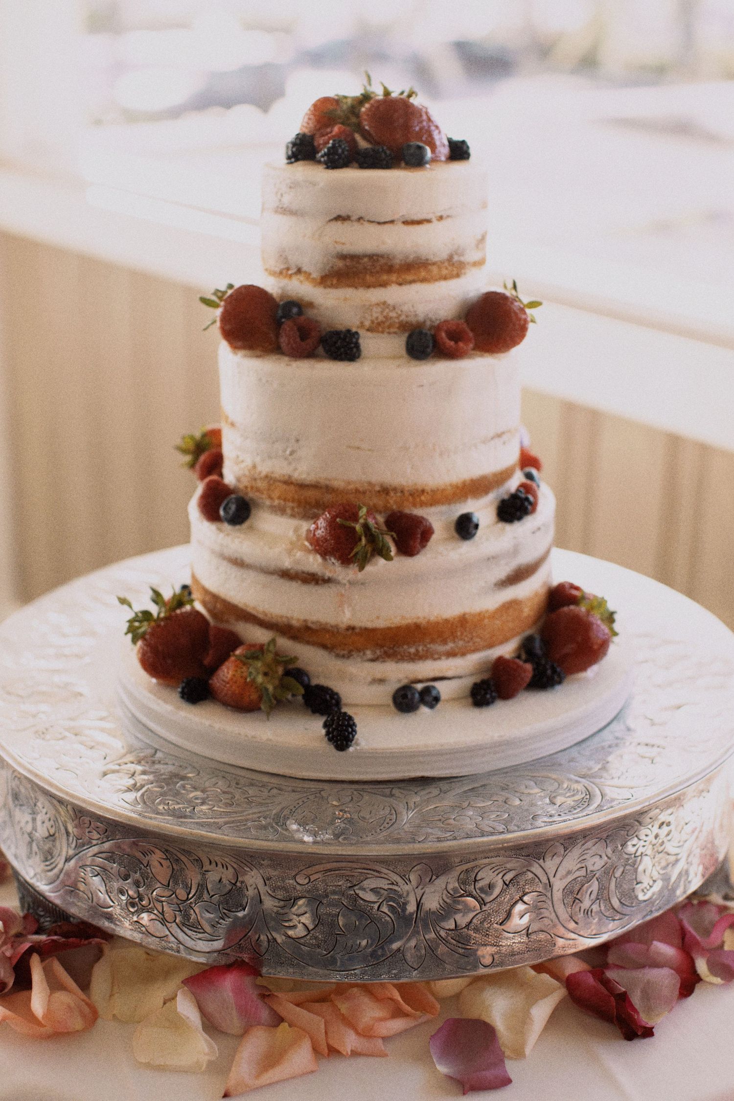 Three-tier semi-naked wedding cake decorated with fresh berries on silver platter.