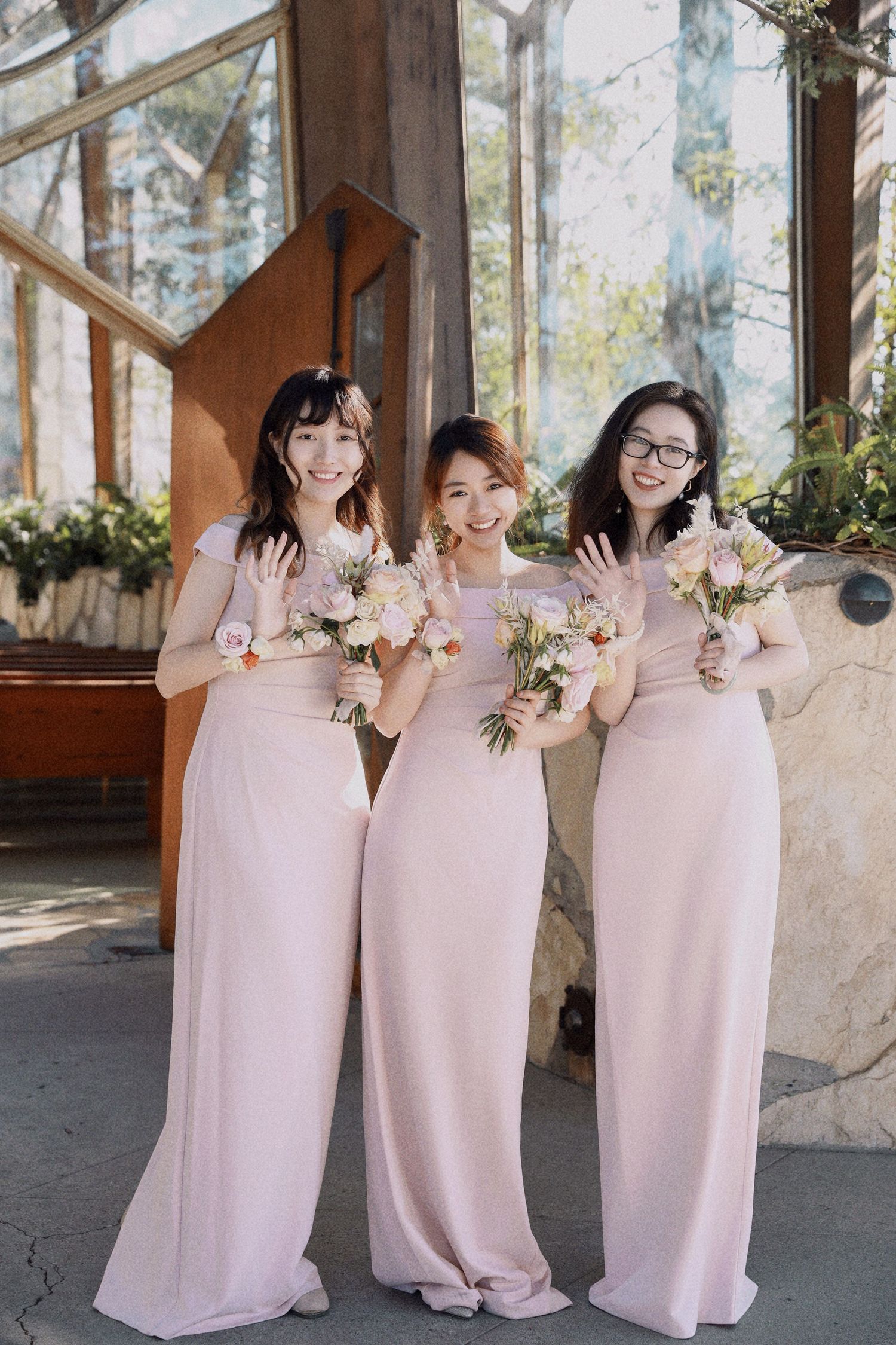 Three bridesmaids in matching pink floor-length dresses hold delicate floral bouquets in an elegant greenhouse setting.