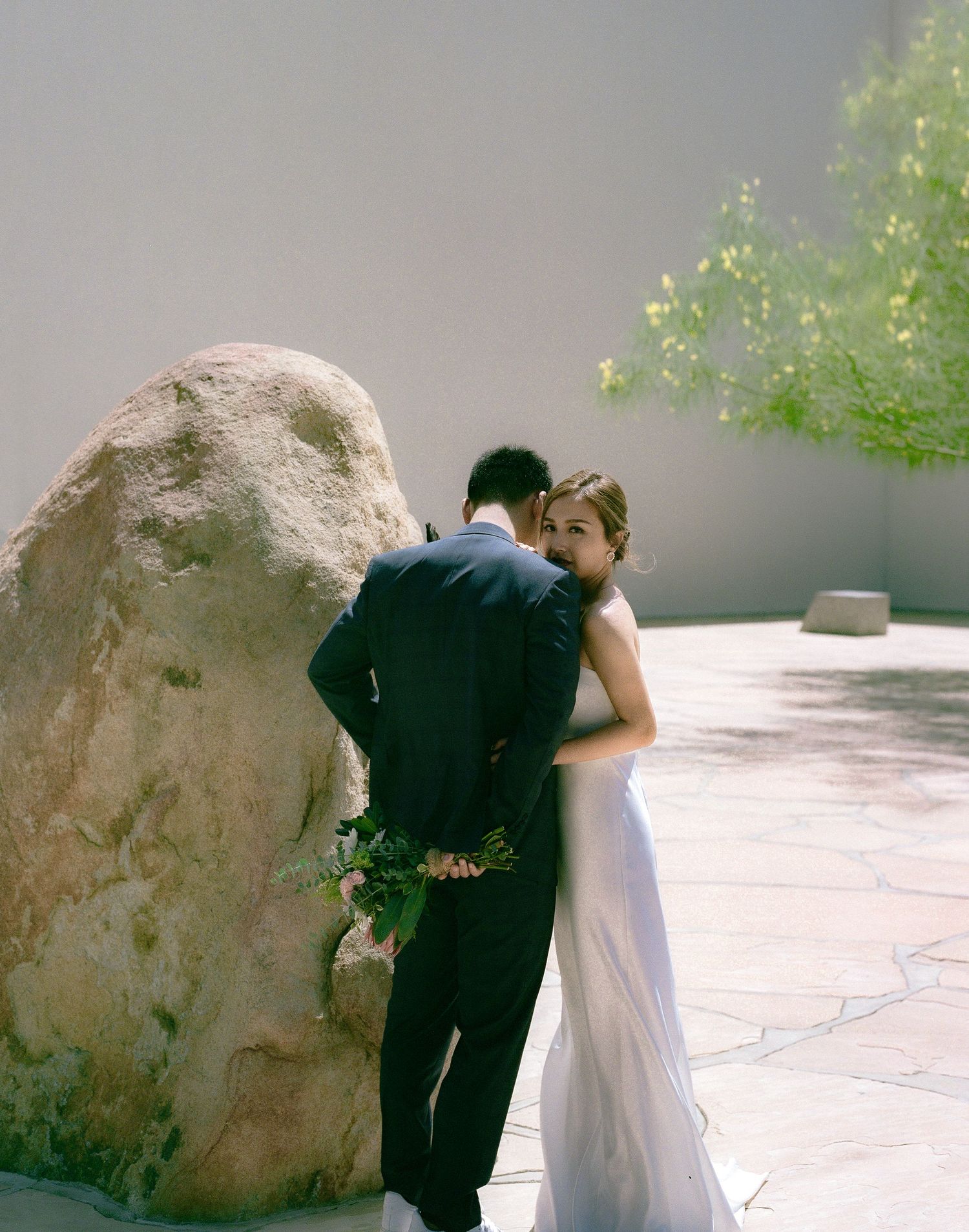 A couple in wedding attire share a tender moment next to a large rock formation.
