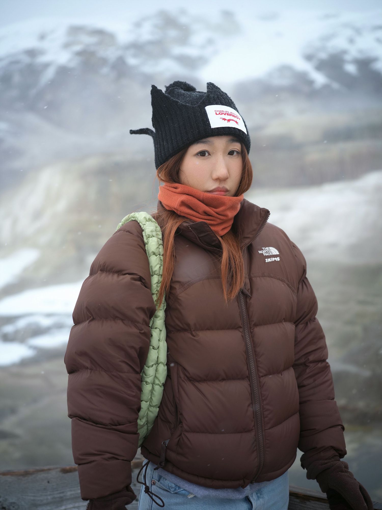 Winter portrait of someone wearing a brown puffer jacket and beanie against a snowy mountain backdrop.