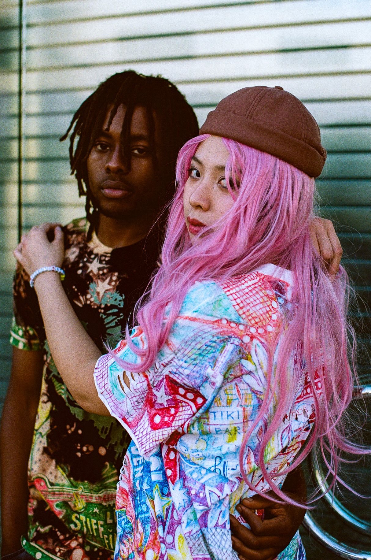 Urban fashion photoshoot featuring contrasting styles with bright pink hair and streetwear against venetian blinds background.