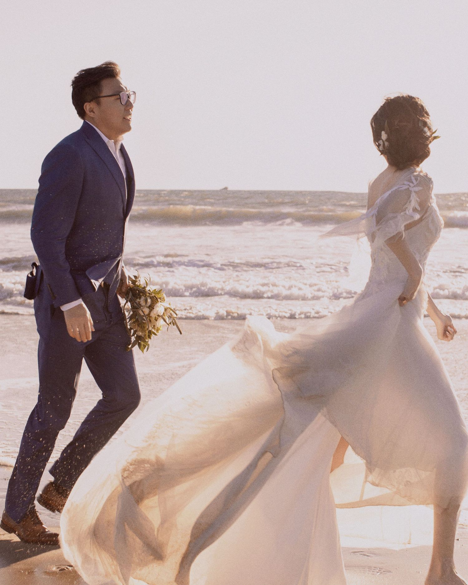 A couple in wedding attire walking on a beach with wind blowing the bride's dress.