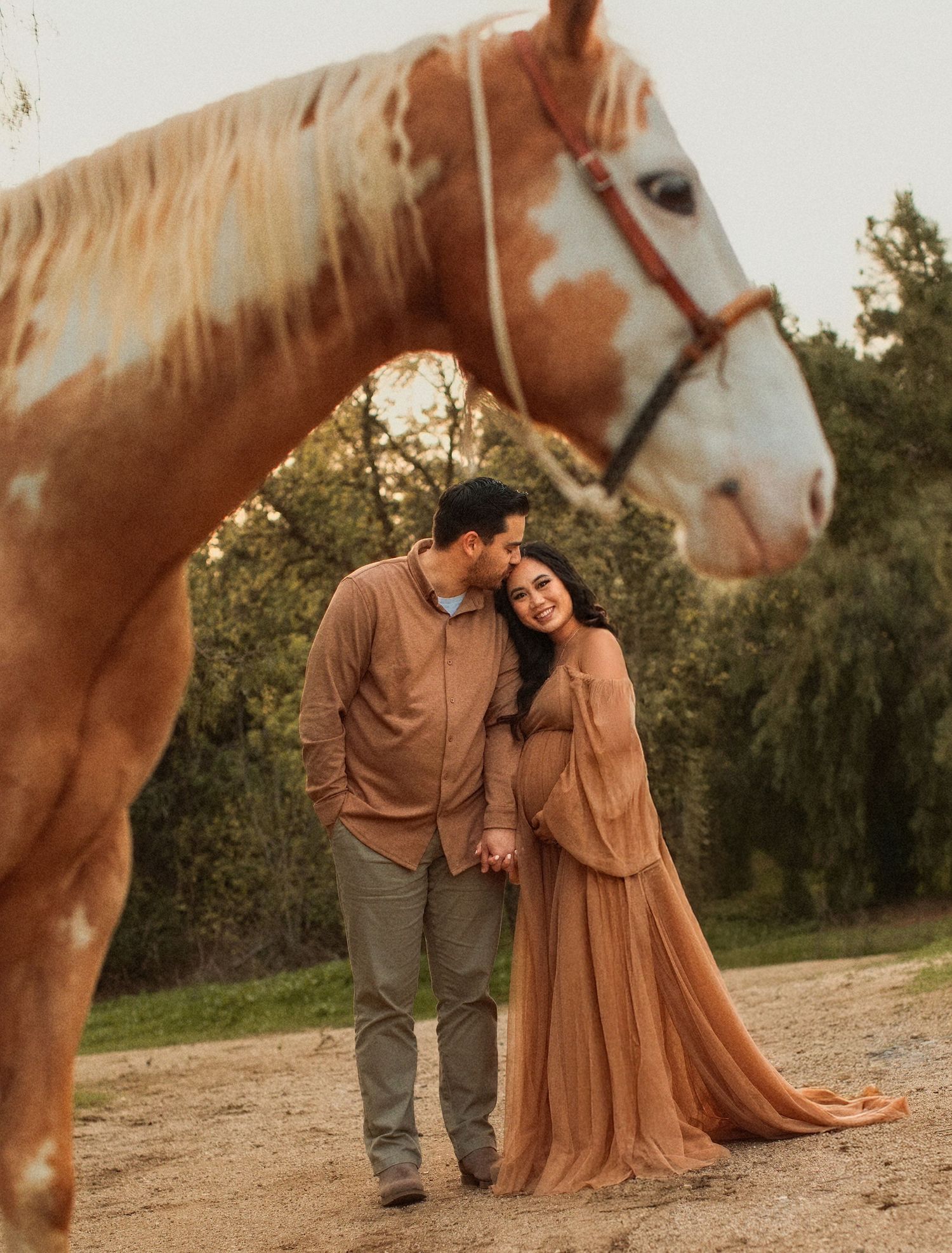 A beautiful brown horse stands near a couple sharing an intimate moment on a dirt path surrounded by greenery.