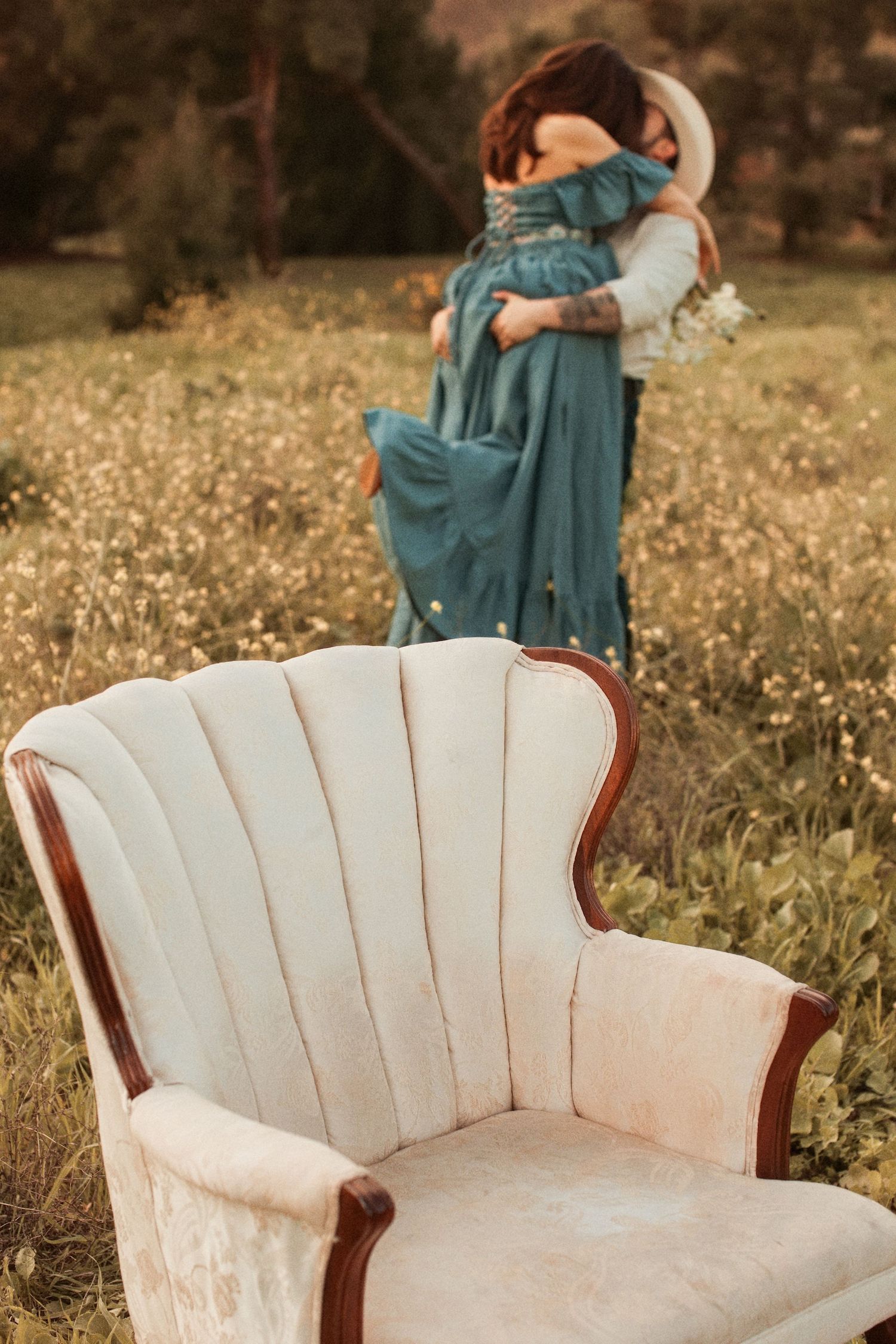 A vintage beige velvet armchair sits in a wildflower meadow with two people embracing in the background.