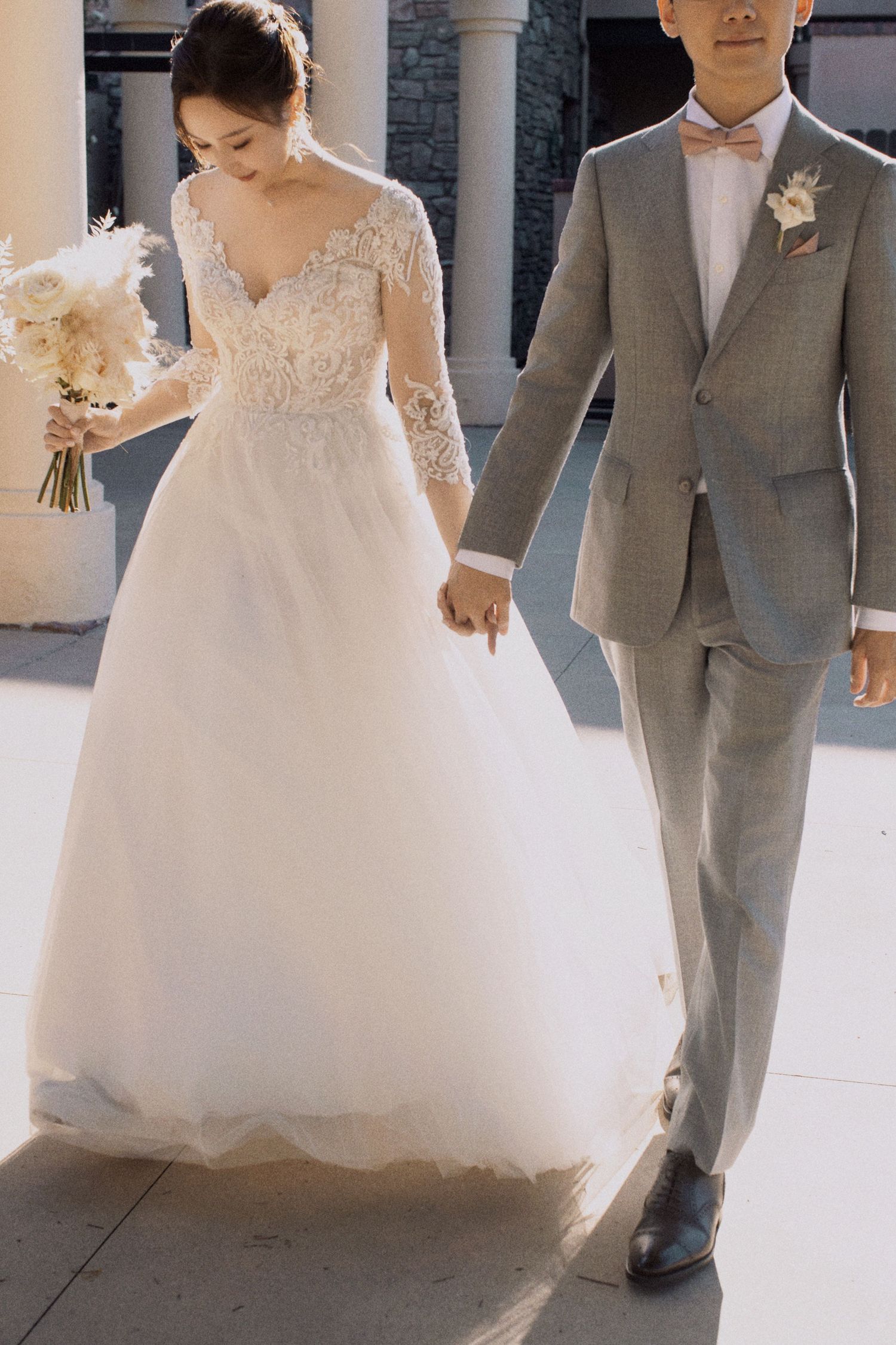 Wedding couple holding hands while walking, bride in lace gown and groom in grey suit.