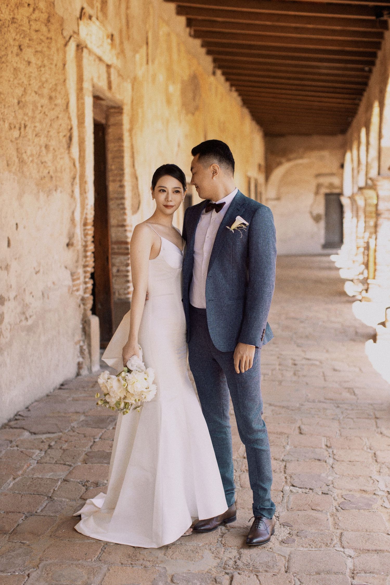 Wedding couple poses in formal attire along historic stone colonnade.
