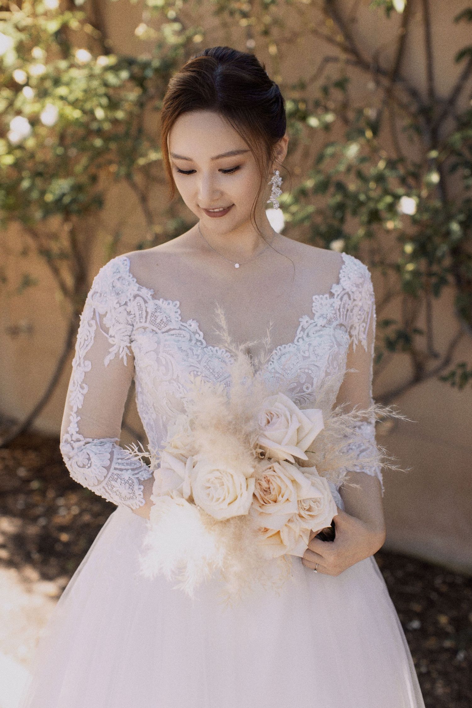 Bride in long-sleeve lace wedding dress holds cream rose bouquet while smiling.