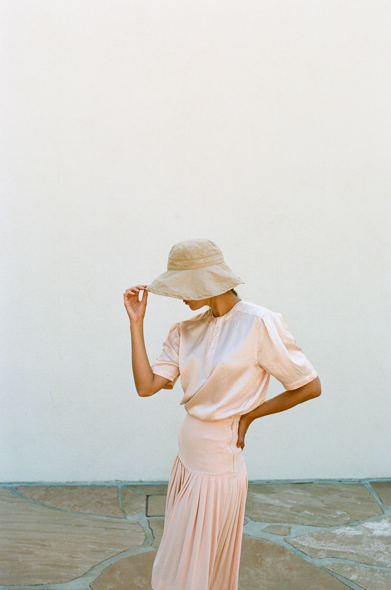 Elegant pose in flowing peach dress and sun hat against light background.
