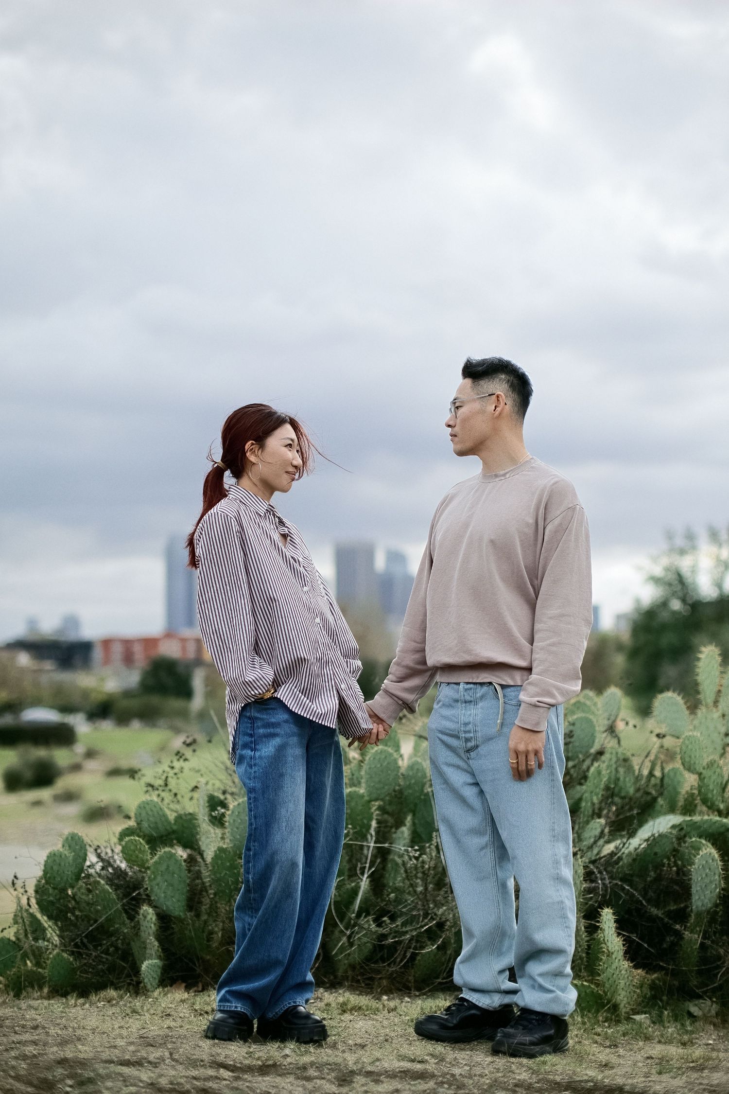 Couple in casual outfits sharing a moment among cacti with city skyline visible.