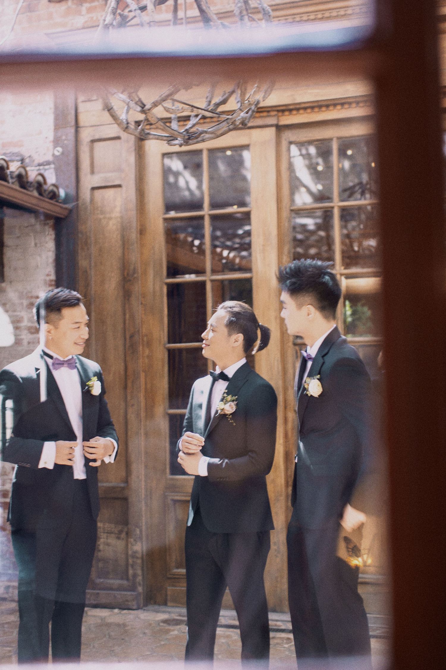 Groomsmen in black tuxedos chatting outside a rustic wooden building.