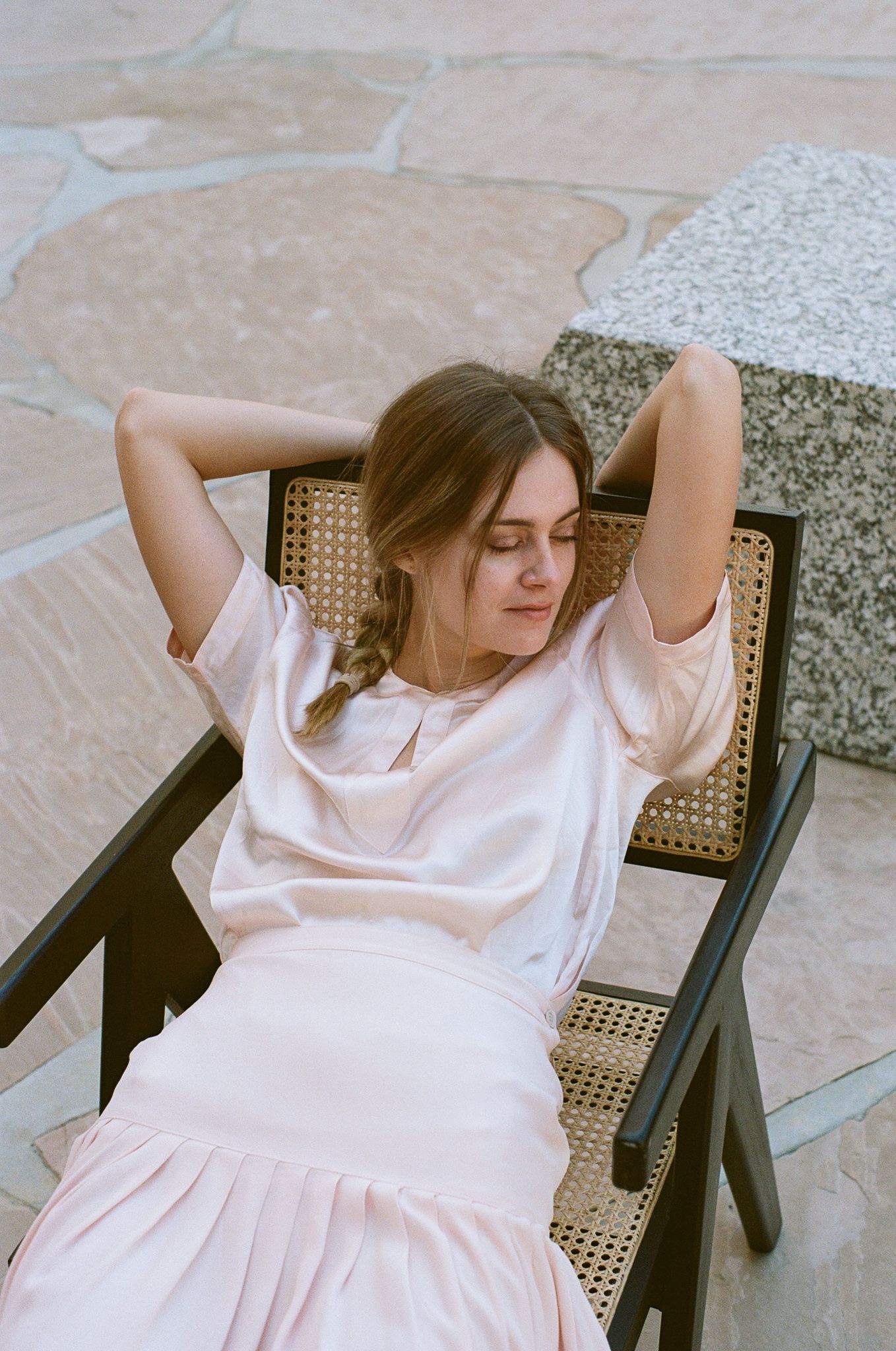 Relaxed pose in cane chair wearing white outfit on stone patio.