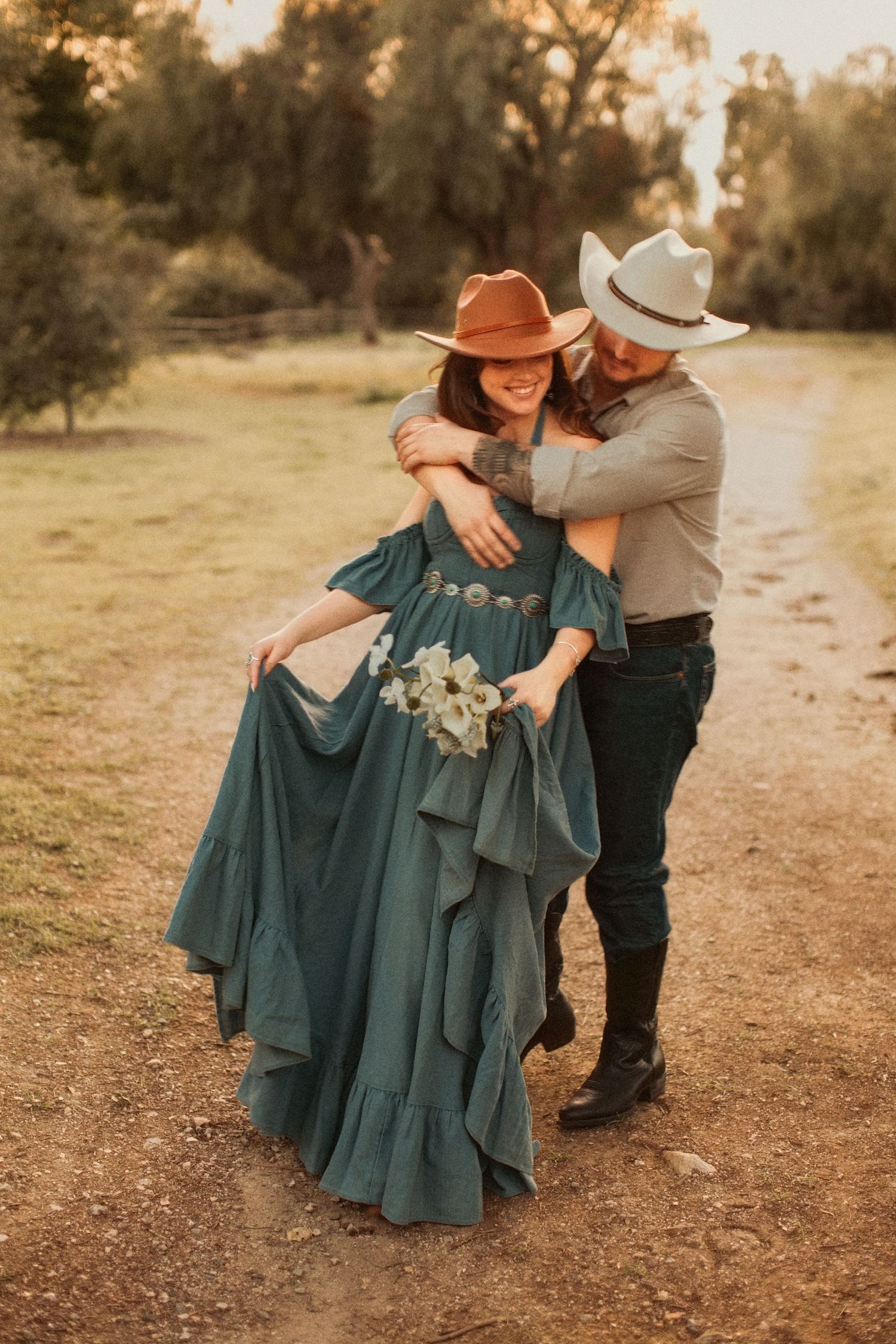 A couple in western attire share an intimate moment on a dirt path at sunset, her teal dress flowing in the golden light.