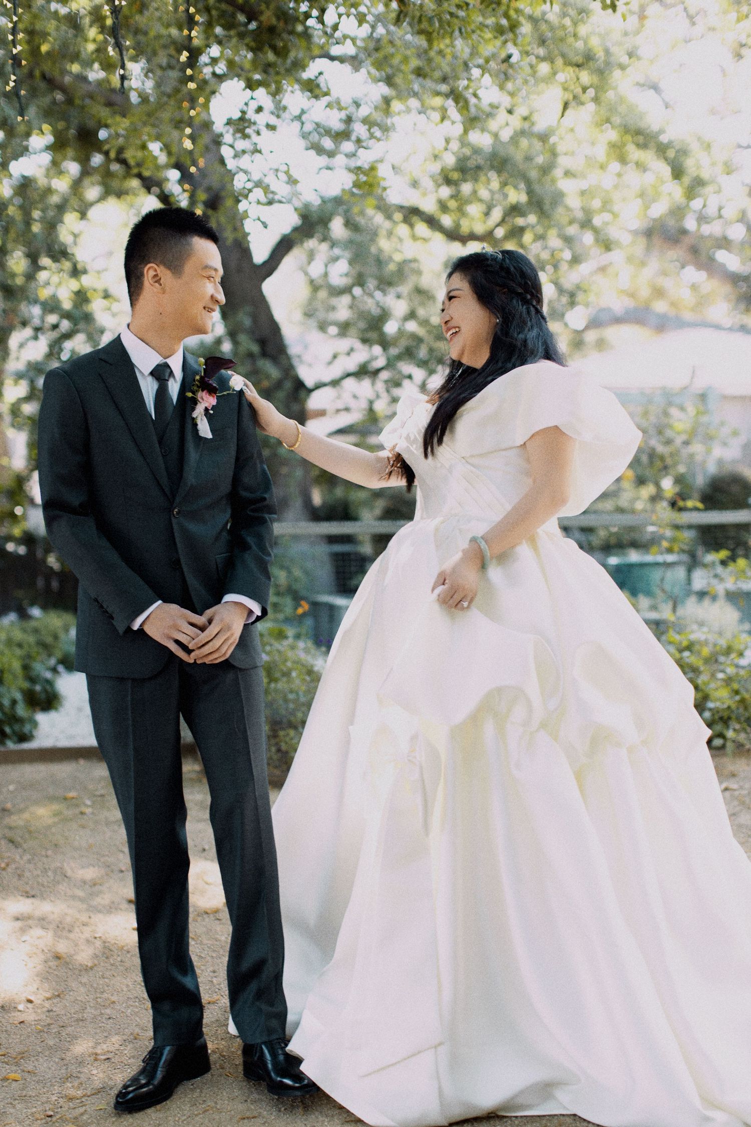 A wedding couple shares a joyful moment outdoors under trees in formal attire.