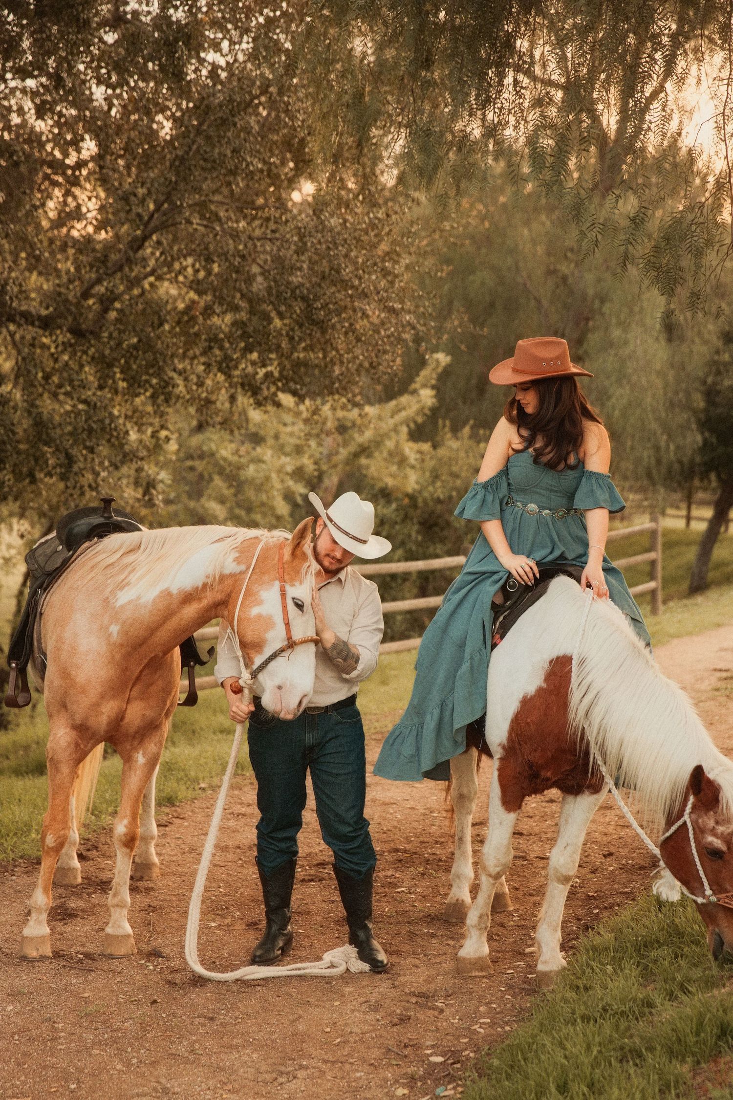 A couple in western attire spend time with their horses on a dirt path during golden hour sunset.