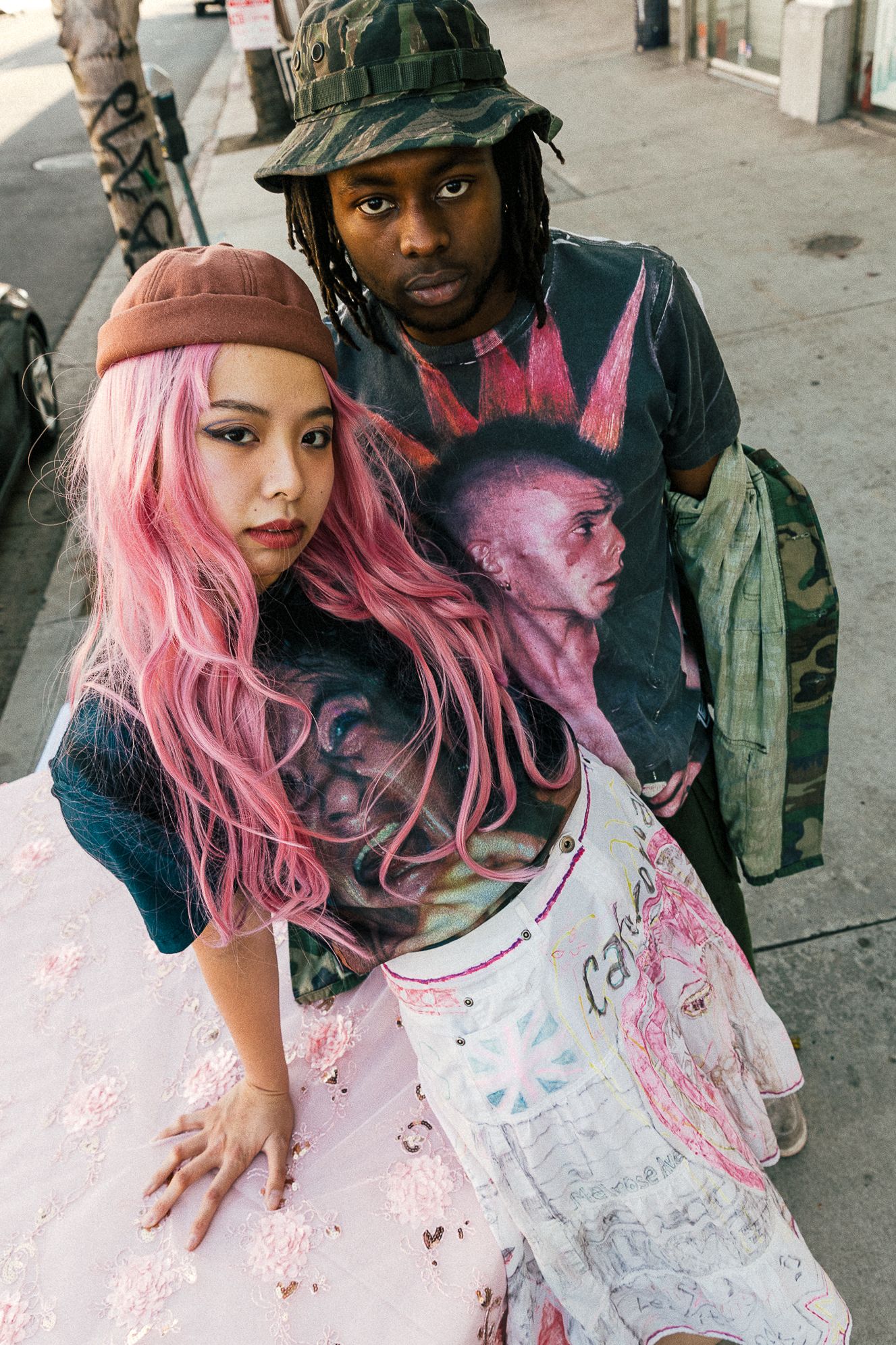 Pink-haired person in white dress and companion with camouflage hat pose artistically on urban street.