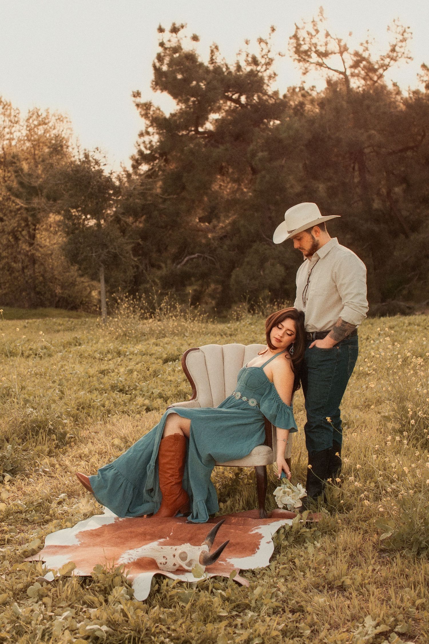 Romantic country couple sitting together on vintage chair in a meadow at sunset with rustic blanket on grass.