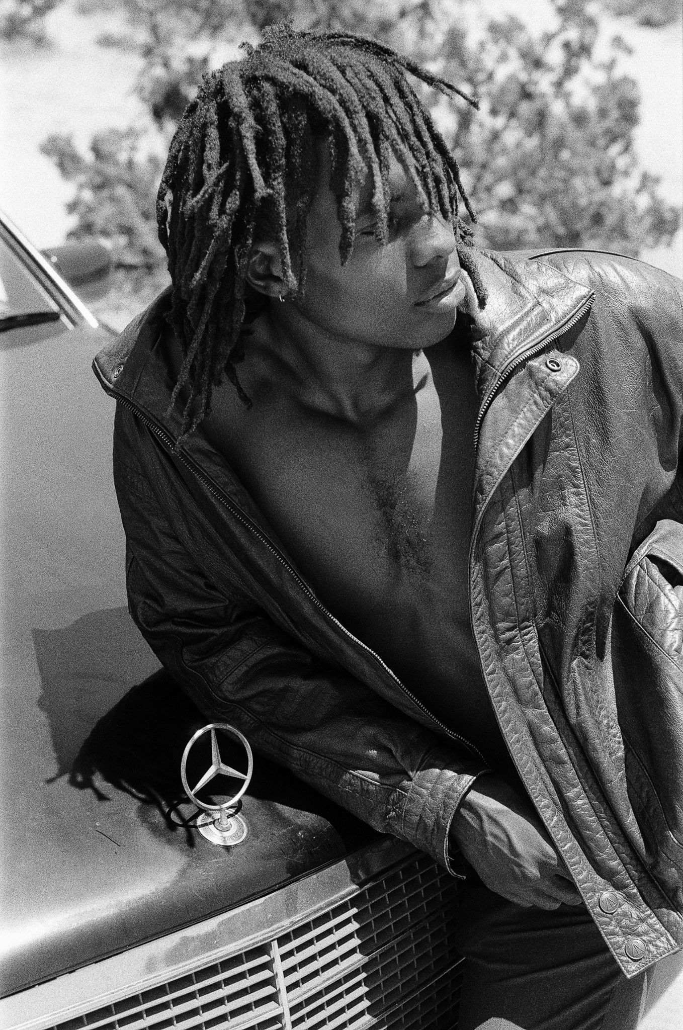 A person in a leather jacket sits on a Mercedes-Benz car with dreadlocks styled hair.