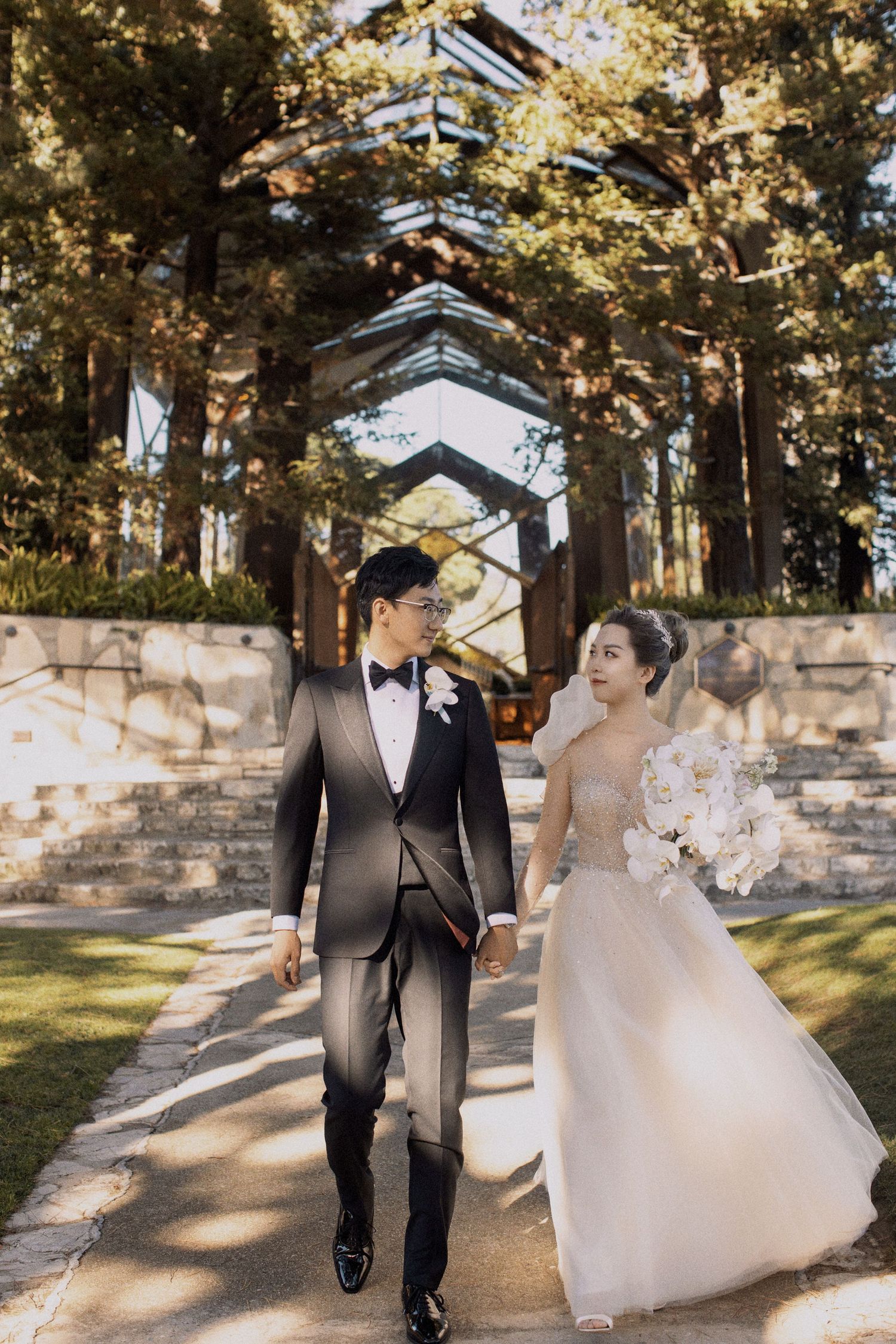A wedding couple walks hand in hand along a tree-lined path with a geometric glass structure behind them.