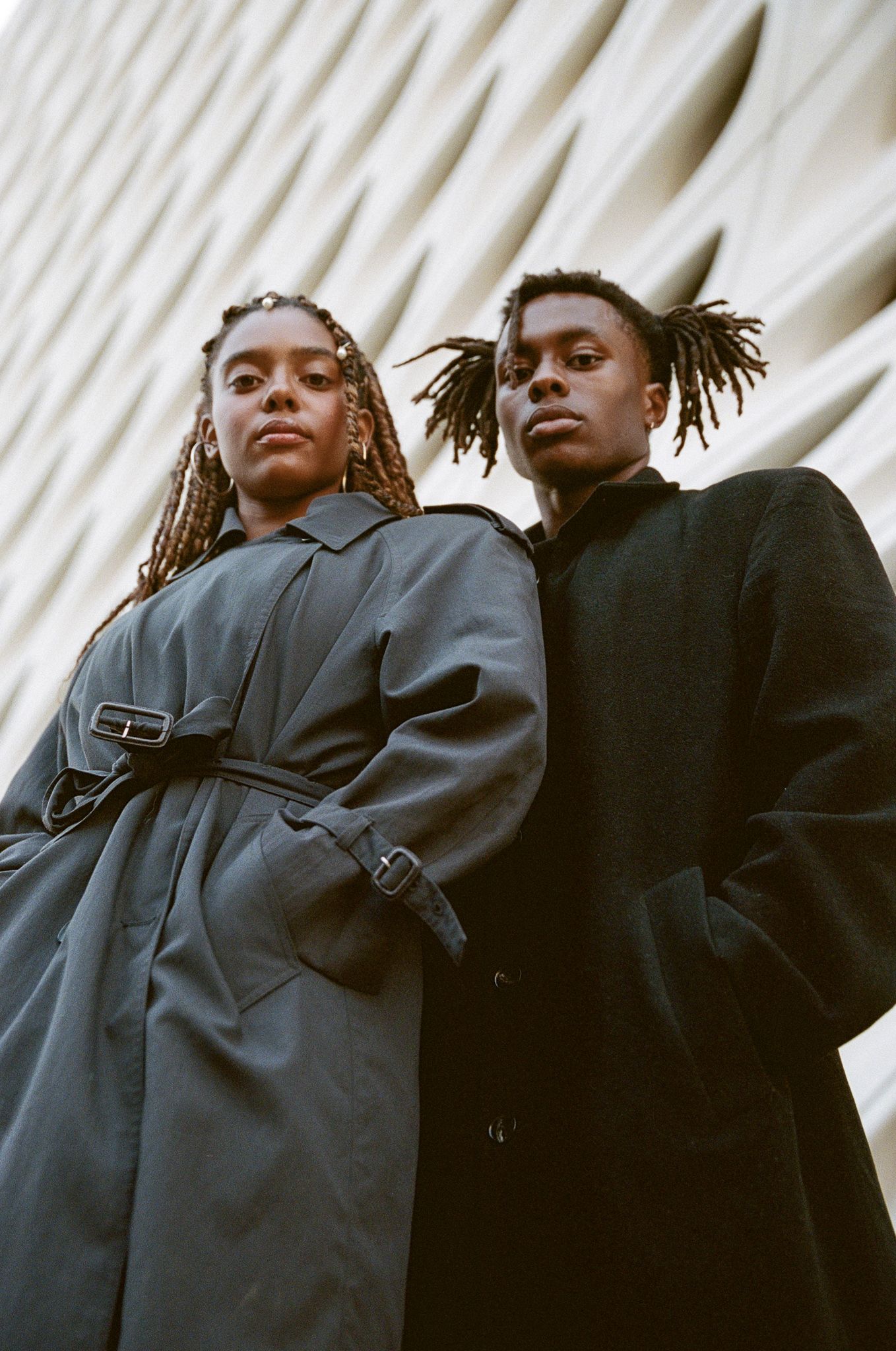 Fashion editorial shot featuring dramatic low angle pose against white architectural background in dark winter coats.