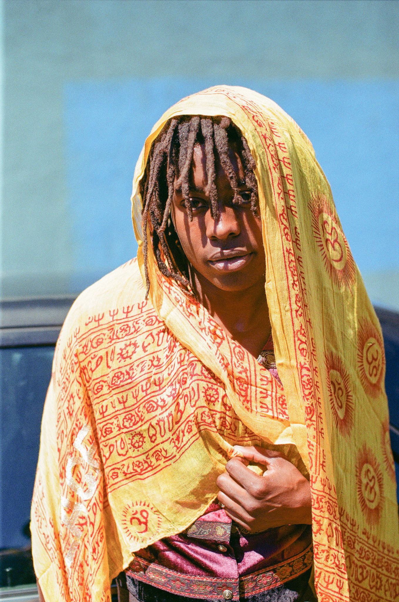 A person wearing a yellow and orange patterned shawl with dreadlocks stands against a blue background.