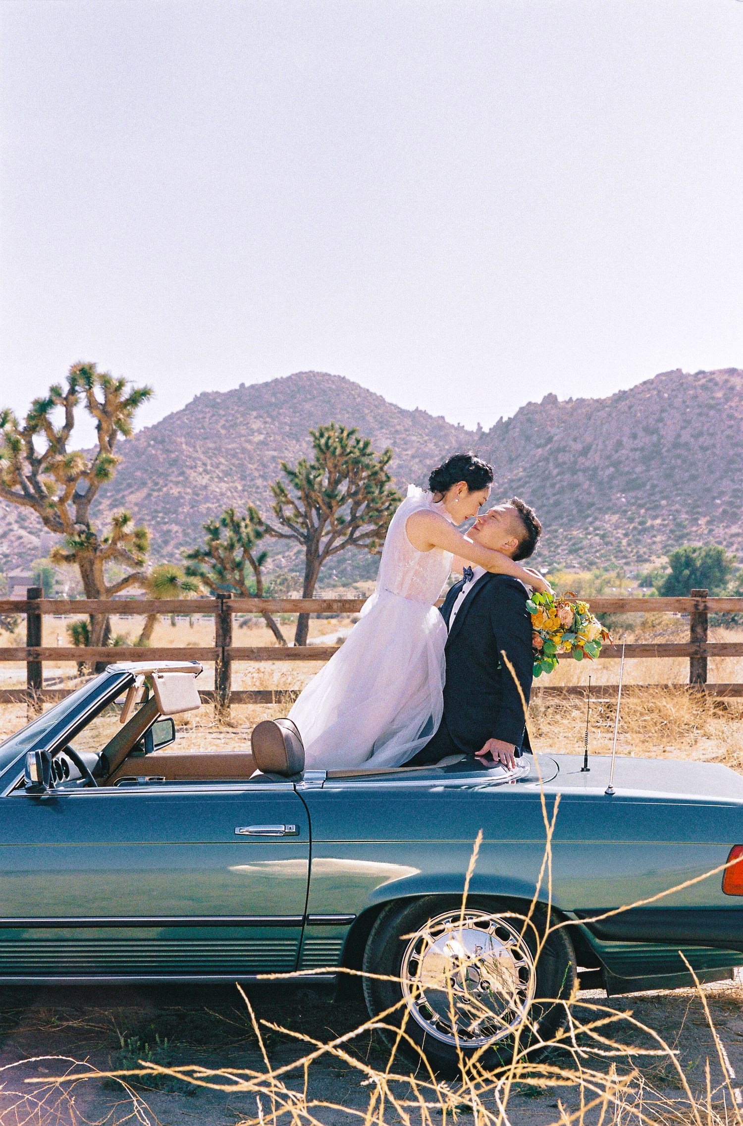 A wedding couple embracing on a classic Mercedes convertible with desert mountains in the background.