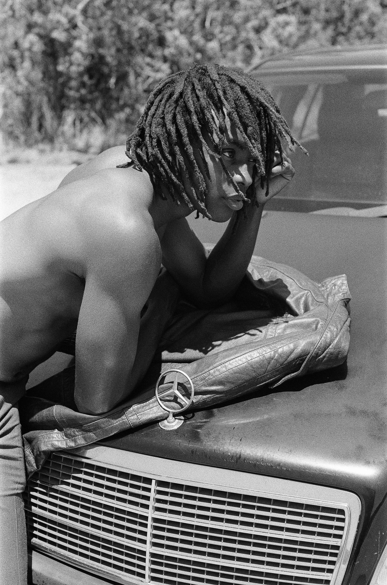 Figure with dreadlocks leaning over a Mercedes-Benz hood in dramatic black and white photography.