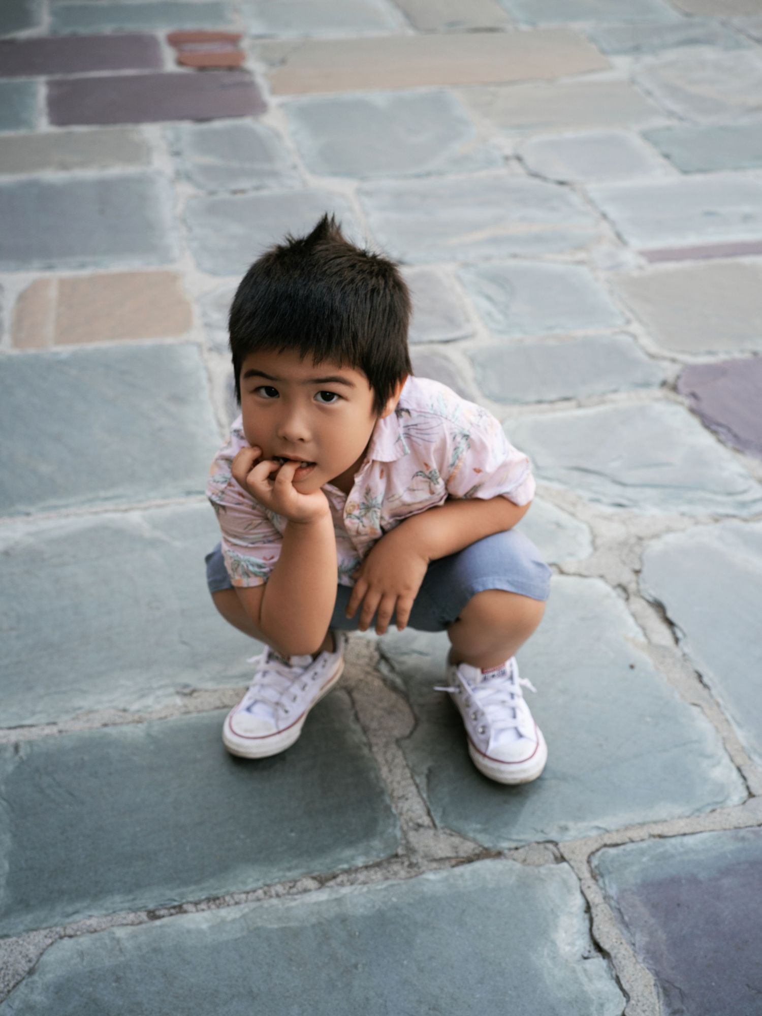 Young child in white sneakers crouching on stone patio looking thoughtful.