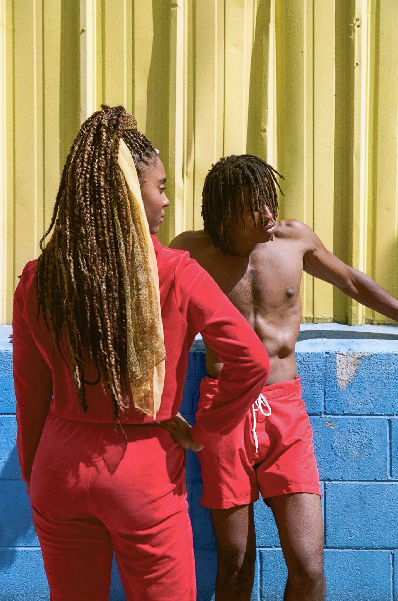 Two people wearing bright red outfits pose against a yellow corrugated wall and blue painted block wall.