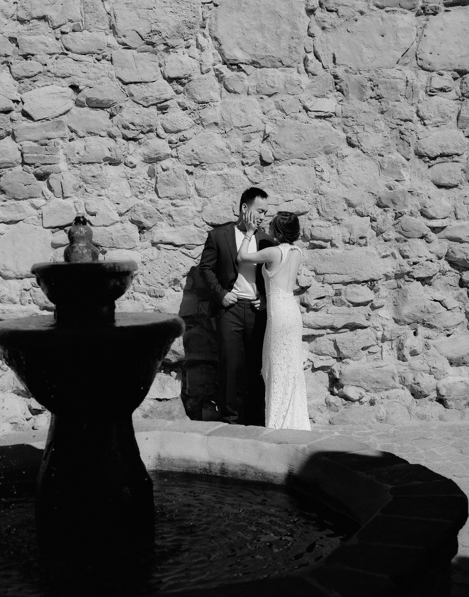 A black and white wedding photo of a couple by a stone wall with a fountain shadow.