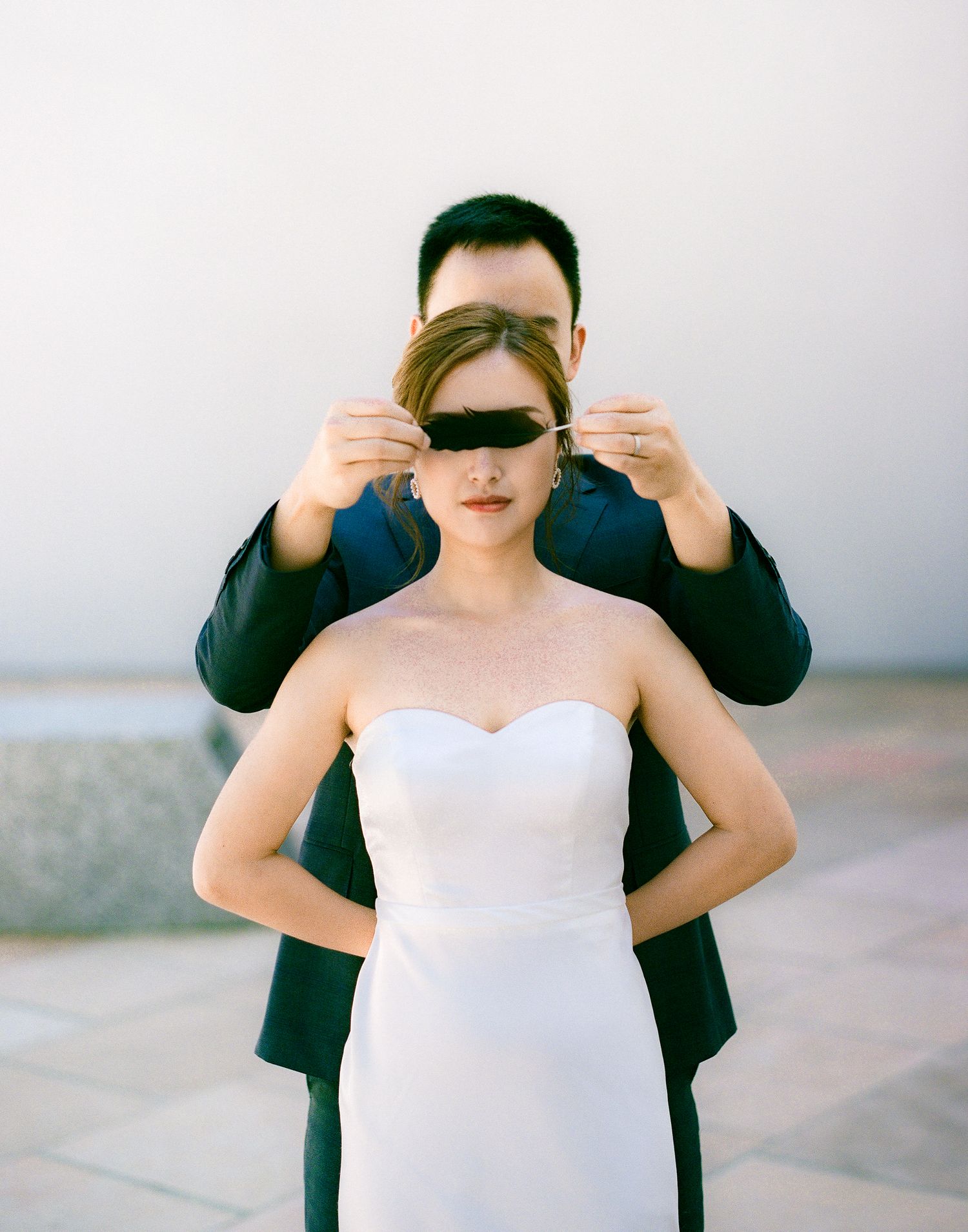 A romantic wedding photo moment with hands covering eyes while wearing a white dress and black jacket.