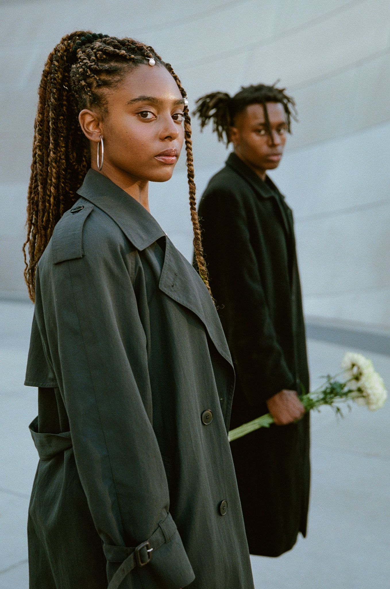 Two people in dark coats pose dramatically against white backdrop, one holding white flowers.