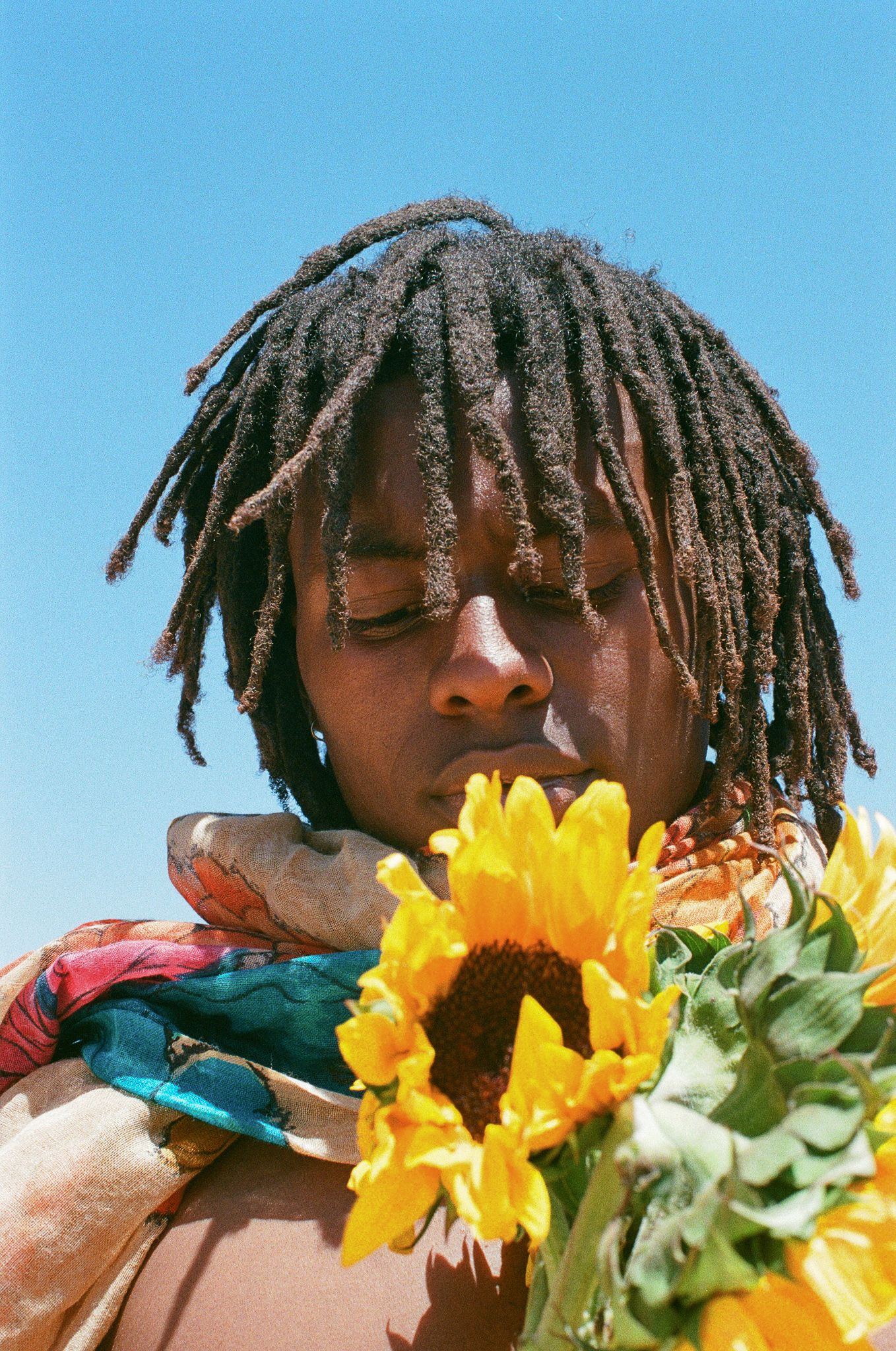 Close-up portrait with dreadlocks holding bright yellow sunflower against blue sky.
