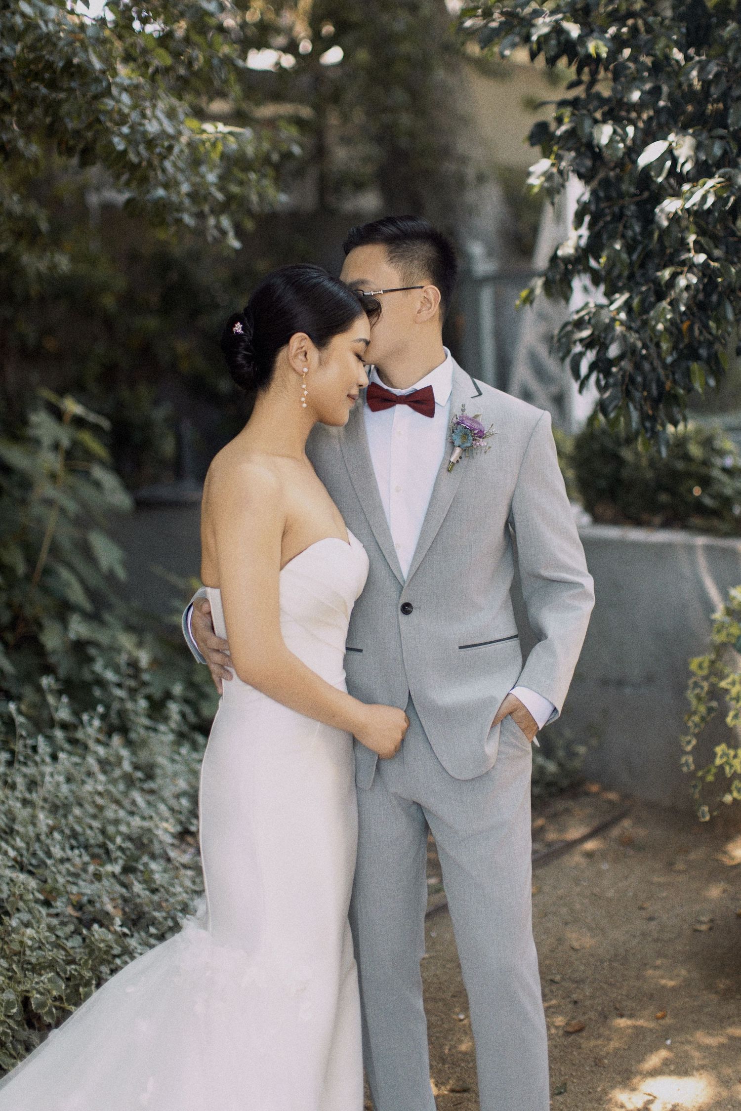 A romantic wedding couple in light gray suit and white dress pose intimately among garden greenery.