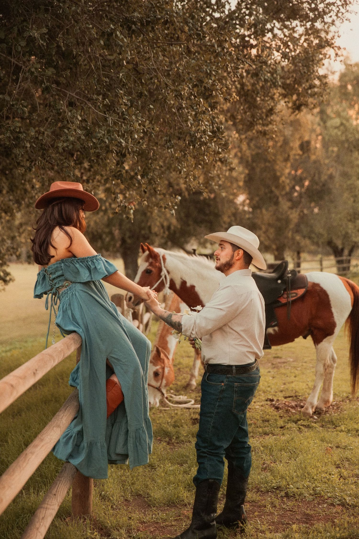 A couple in western attire shares a romantic moment by a wooden fence with horses grazing in the background at sunset.