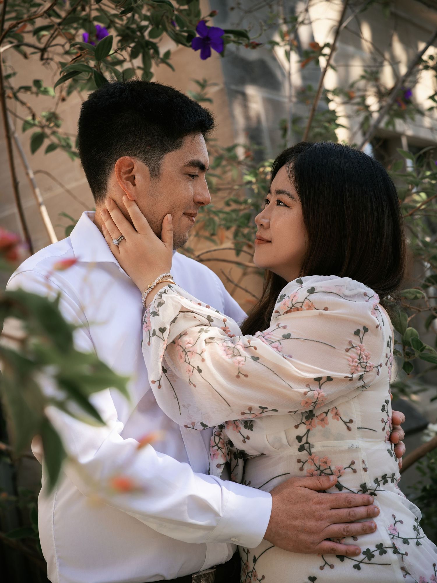 A couple in romantic embrace among flowering vines, wearing matching floral and white outfits in soft natural light.