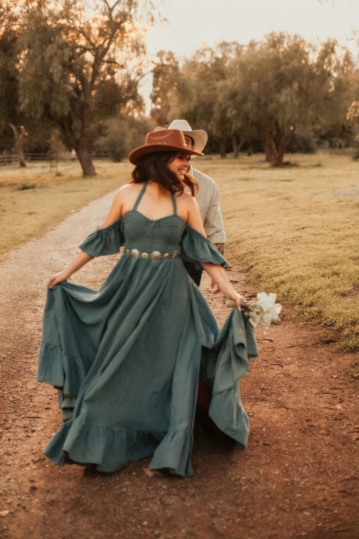 Woman in flowing teal dress and brown hat walks along dirt path during golden sunset in wooded area.