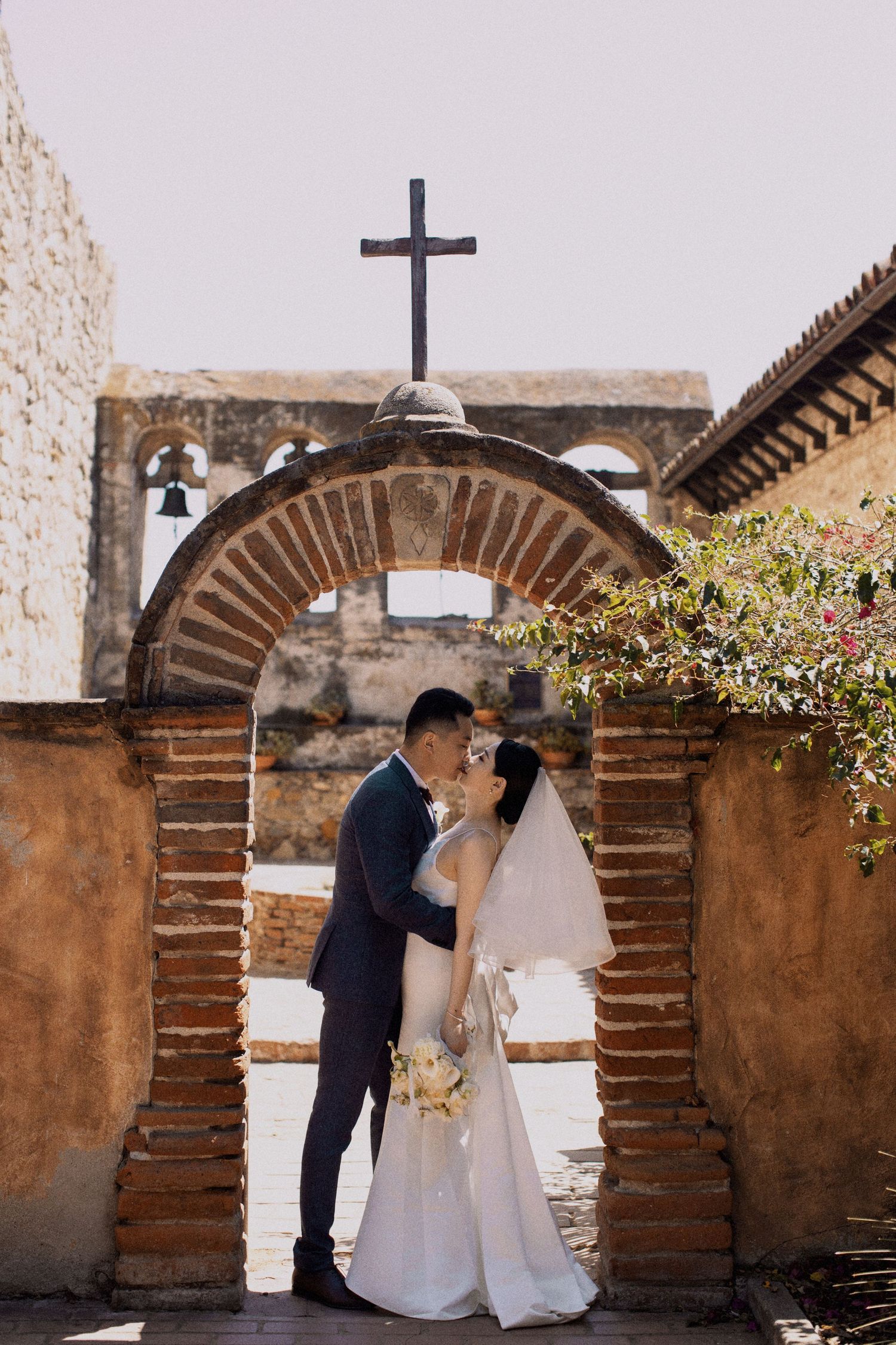 Newlyweds share a romantic moment under a rustic brick archway with a cross.