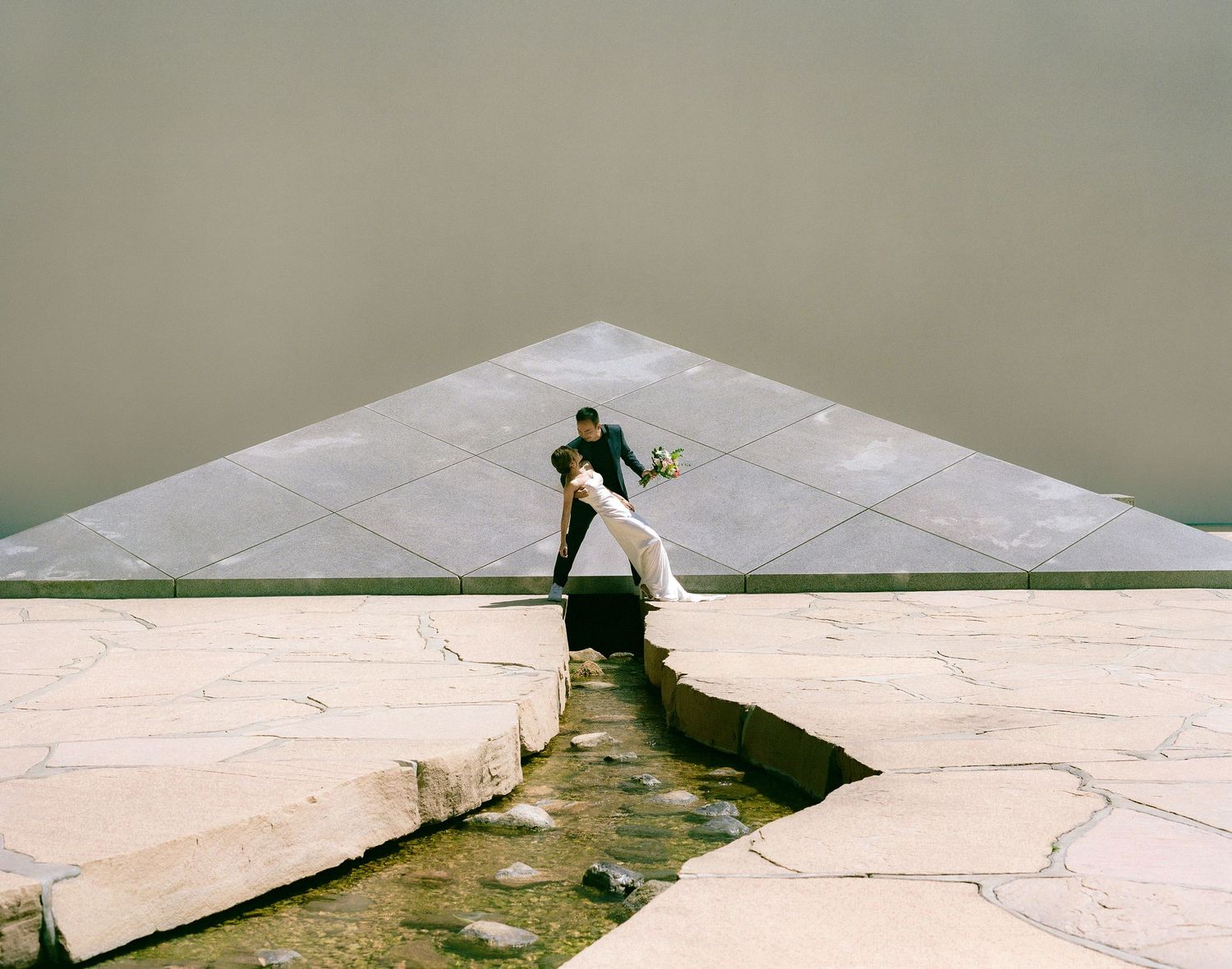 A dramatic architectural shot showing two people crossing over water on geometric stone platforms.