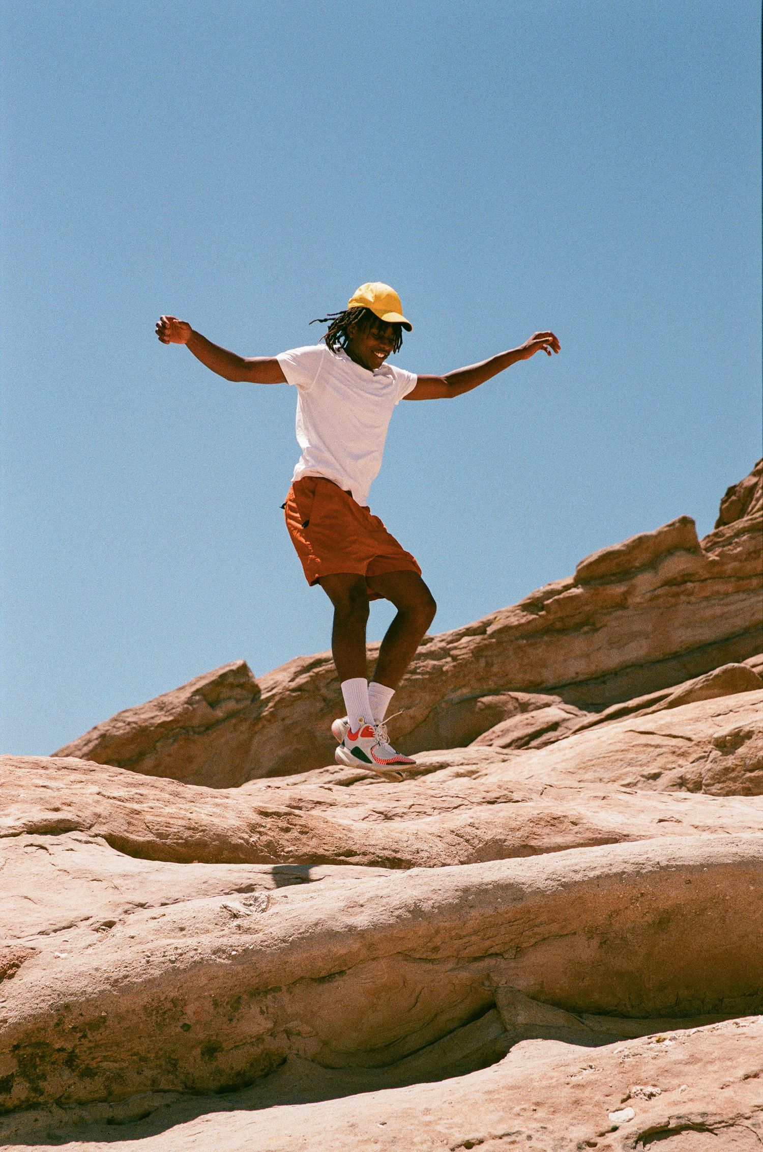 An energetic figure in orange shorts and yellow cap jumps across desert rock formations.