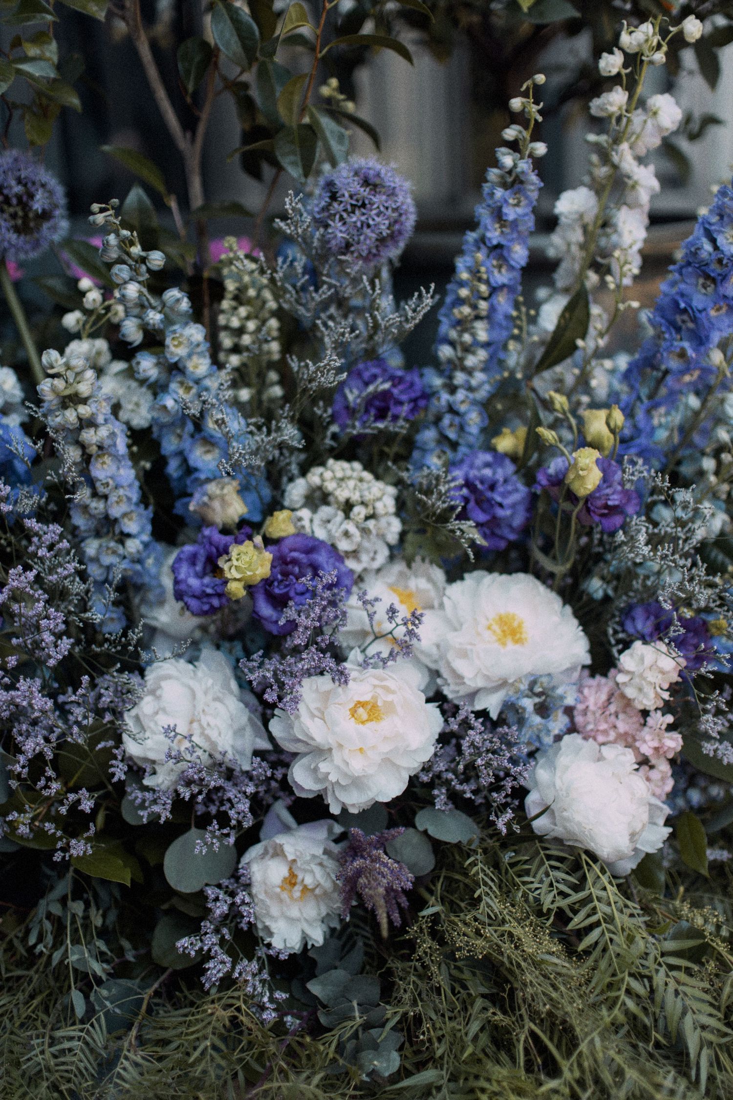 A lush floral arrangement featuring white peonies, blue delphiniums, and purple blooms with greenery.