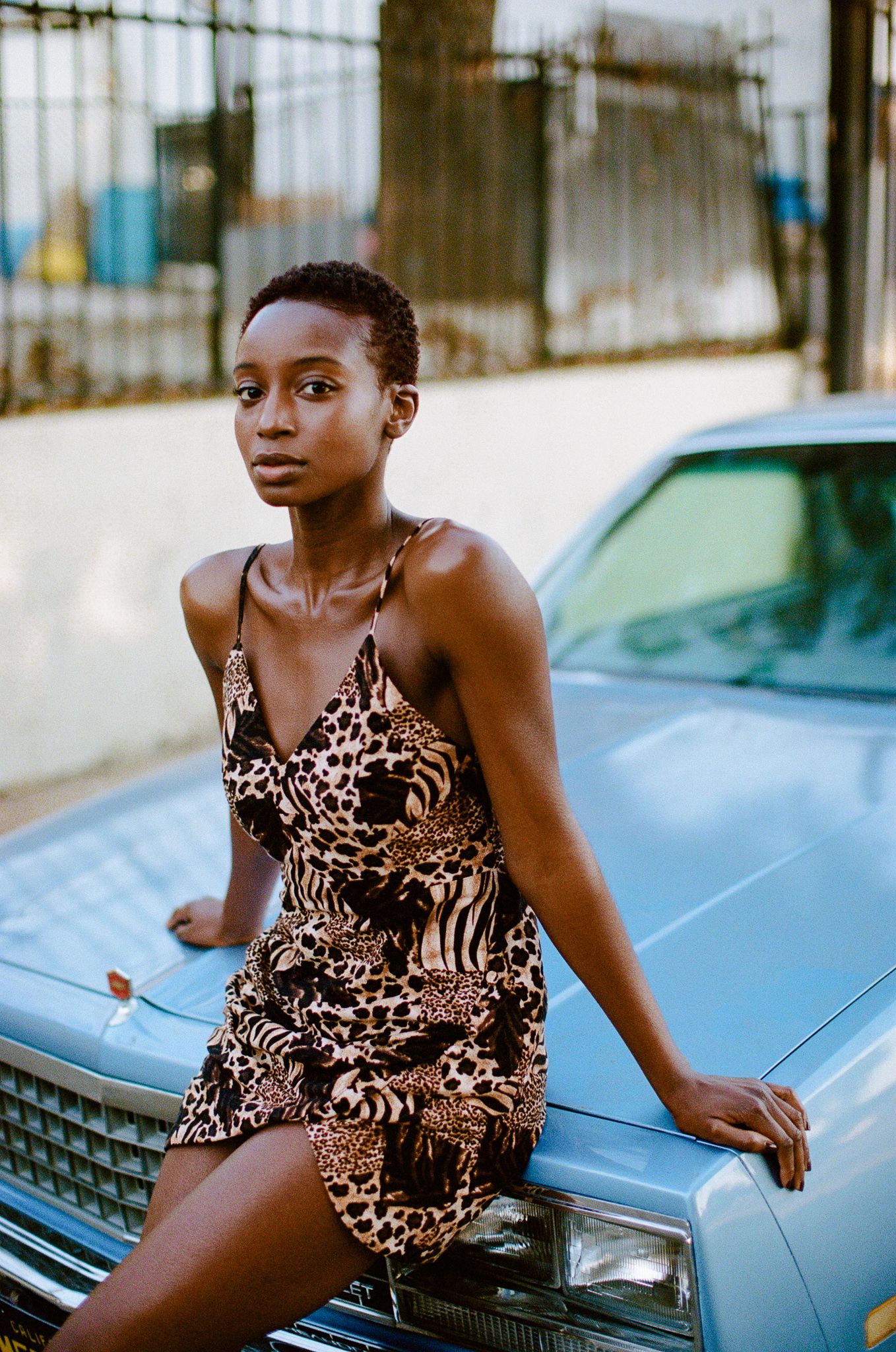 Woman in animal print dress poses on vintage blue car hood.