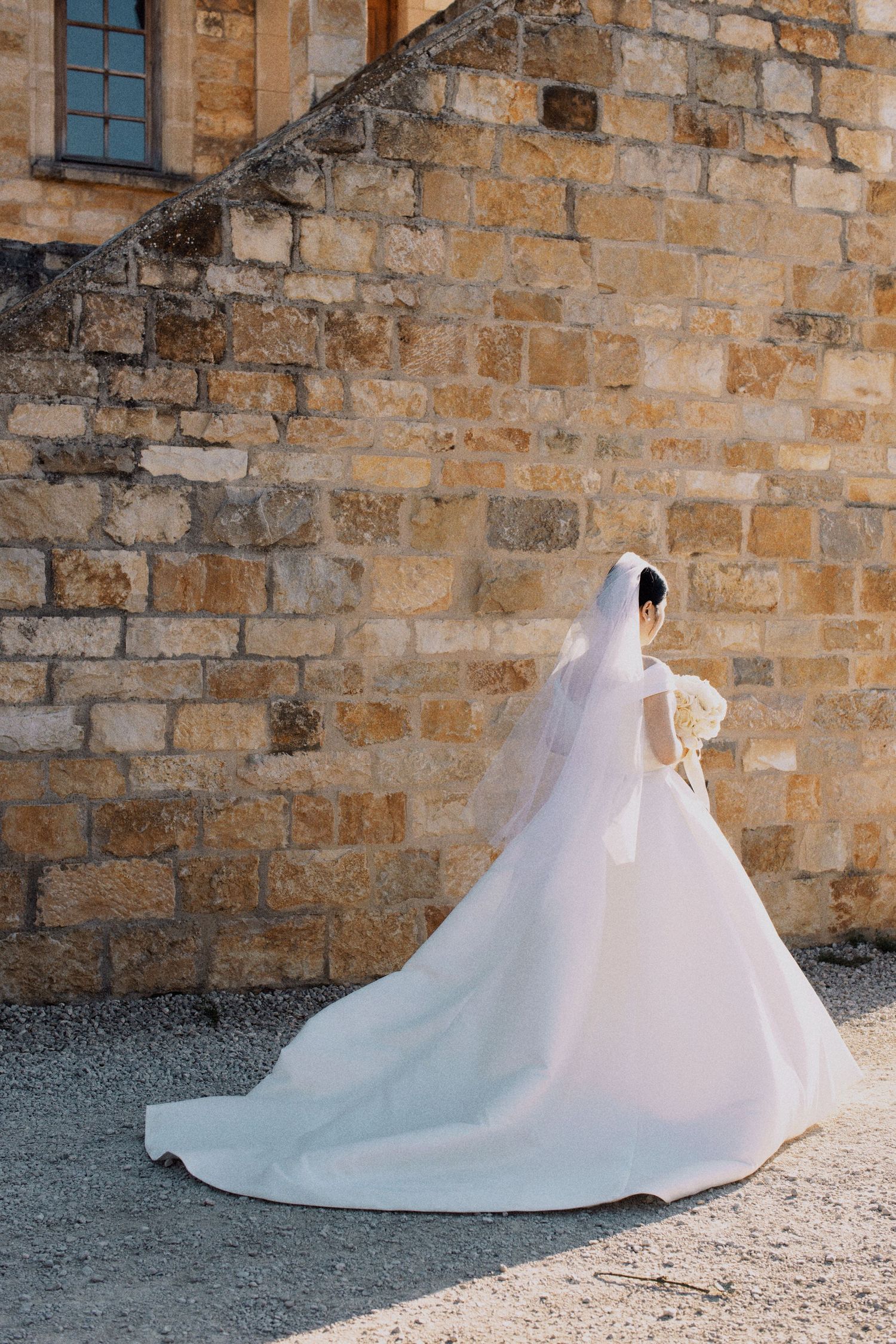 A bride in a flowing white wedding gown stands against a rustic stone wall at a historic venue.