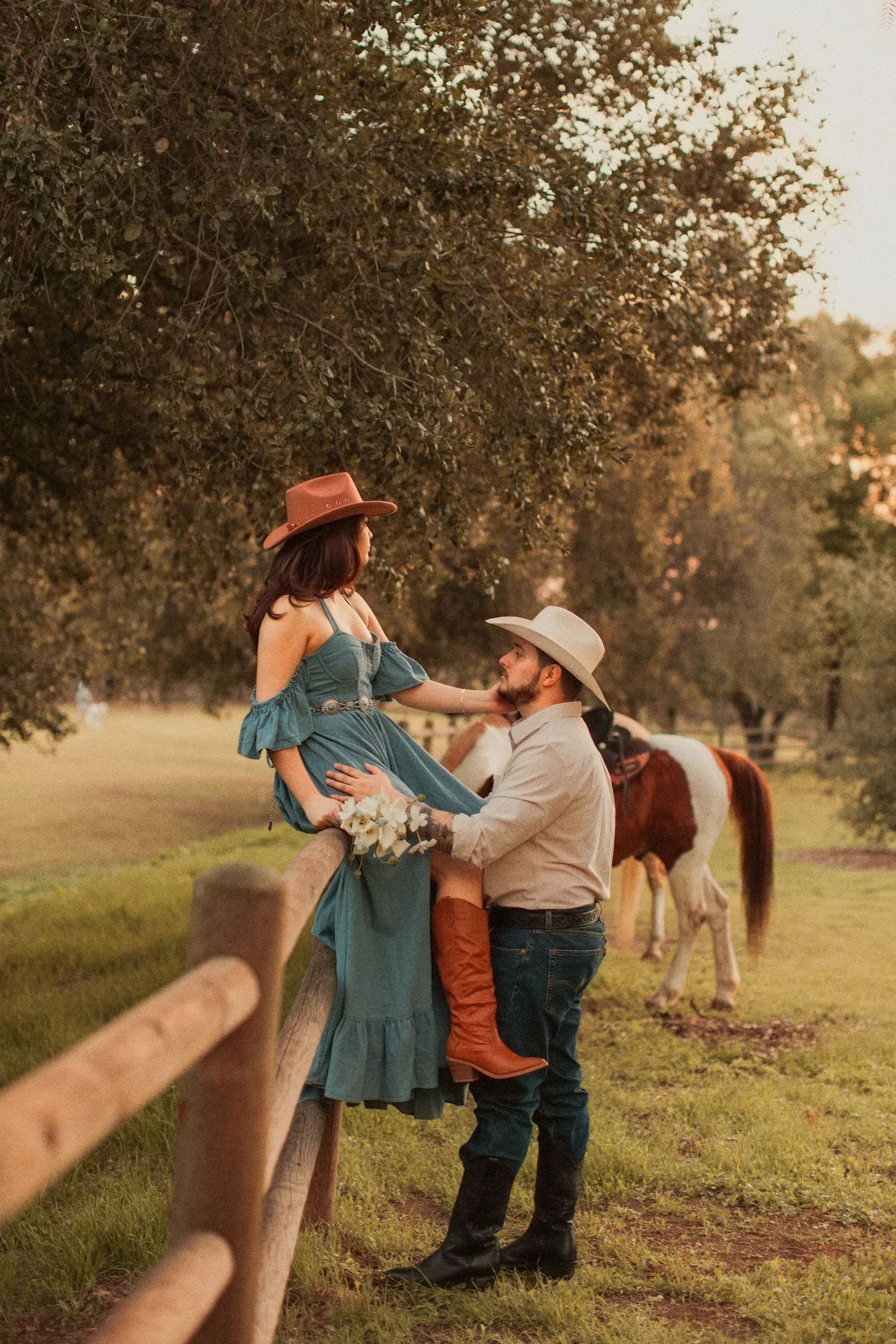 A romantic western-style photo shoot in a field with a couple in cowboy attire and a horse in the background.