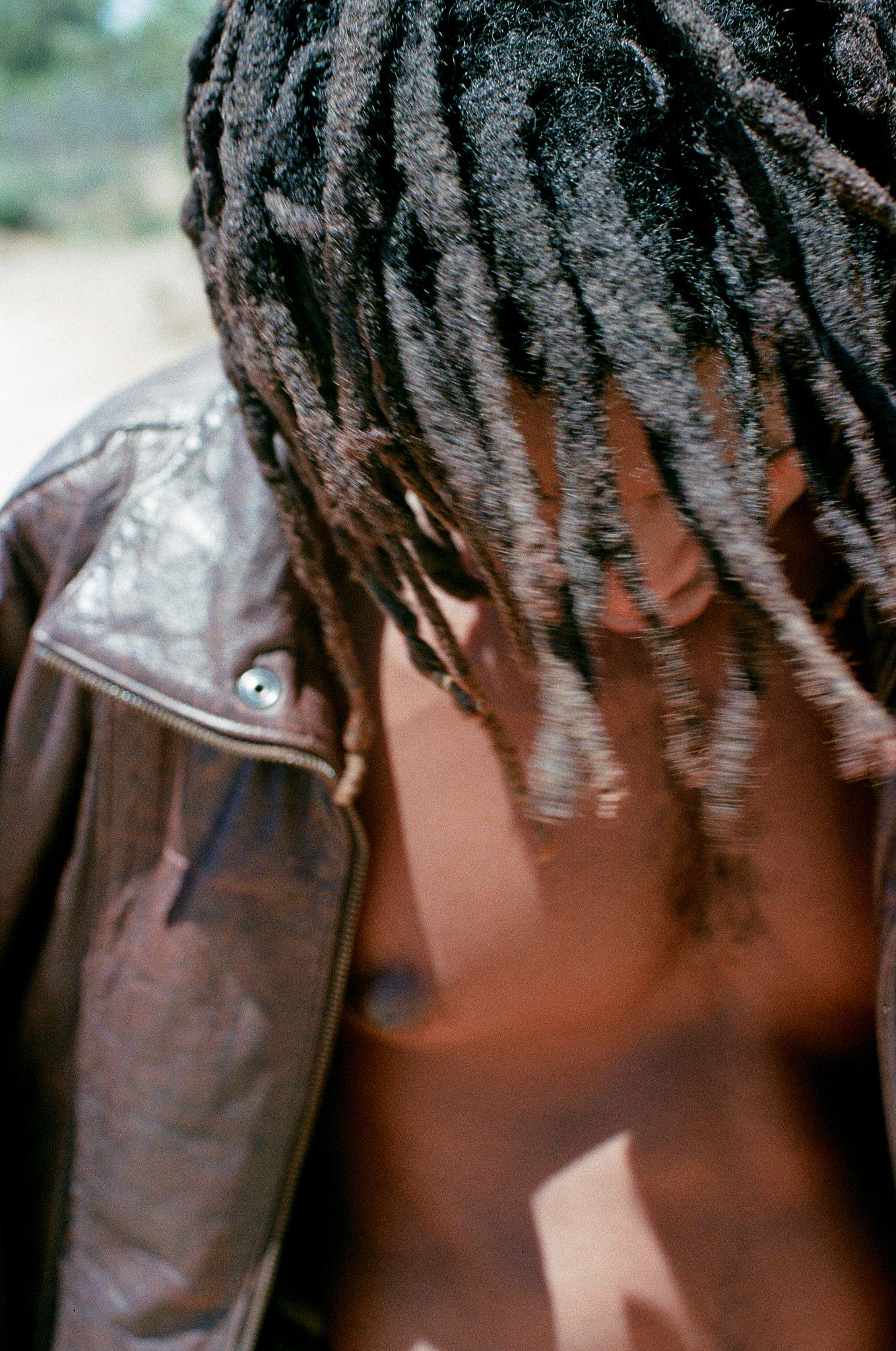Close-up portrait showing dreadlocks and brown leather jacket against blurred background.