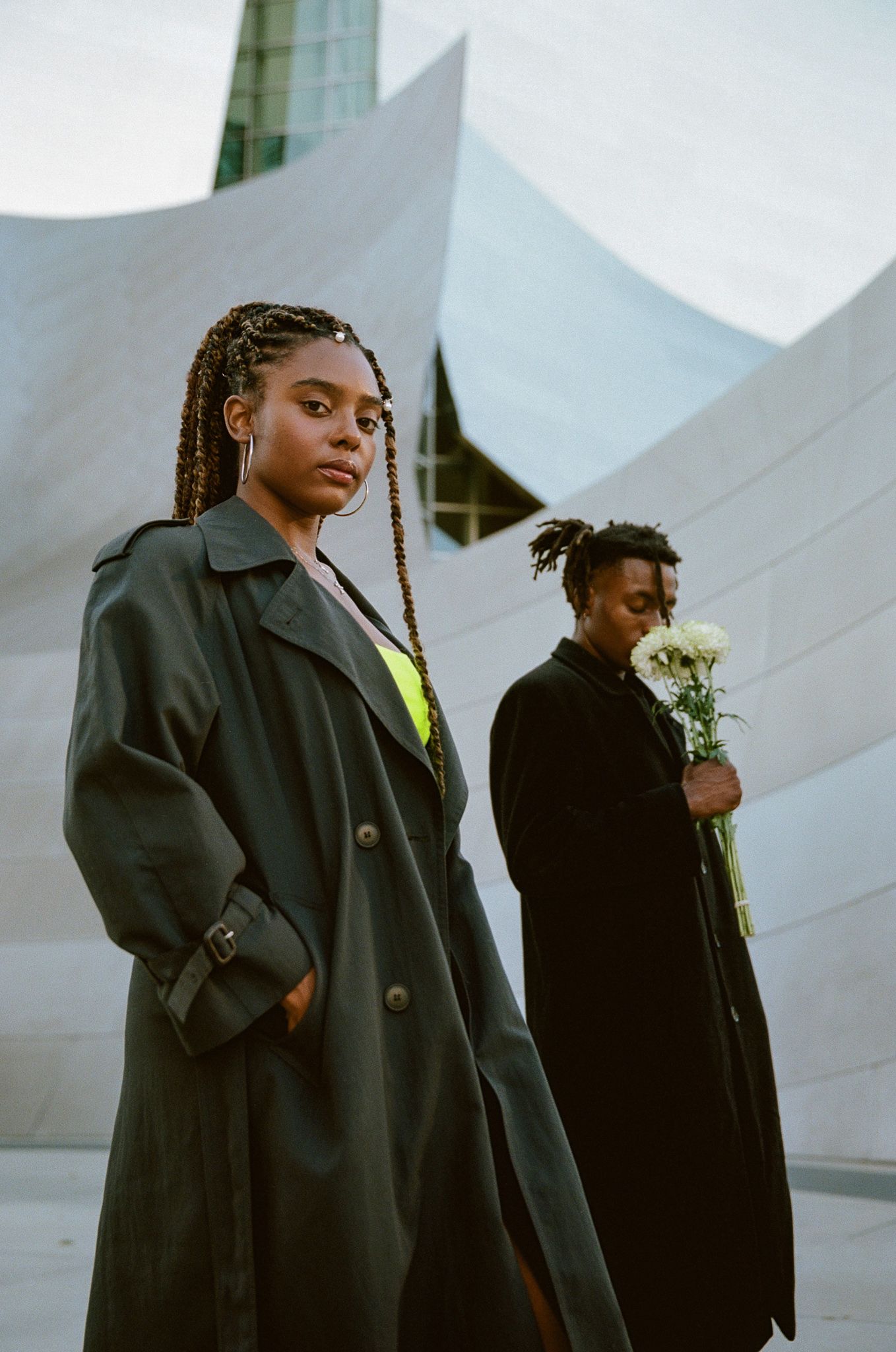 Two people in long black coats pose against a modern architectural backdrop with white geometric shapes.