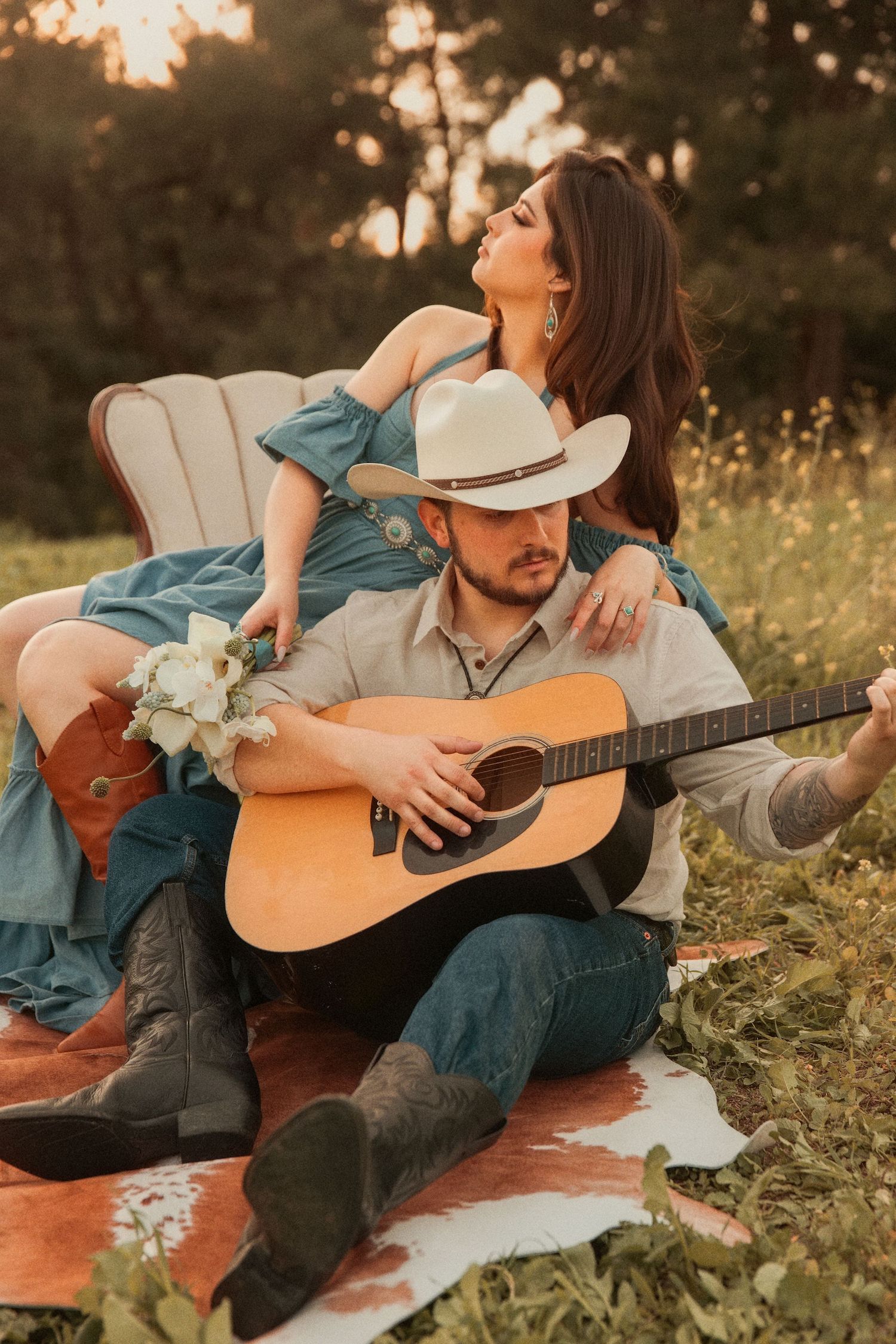 Romantic country couple sitting on blanket at sunset with guitar and flowers in rustic outdoor setting.