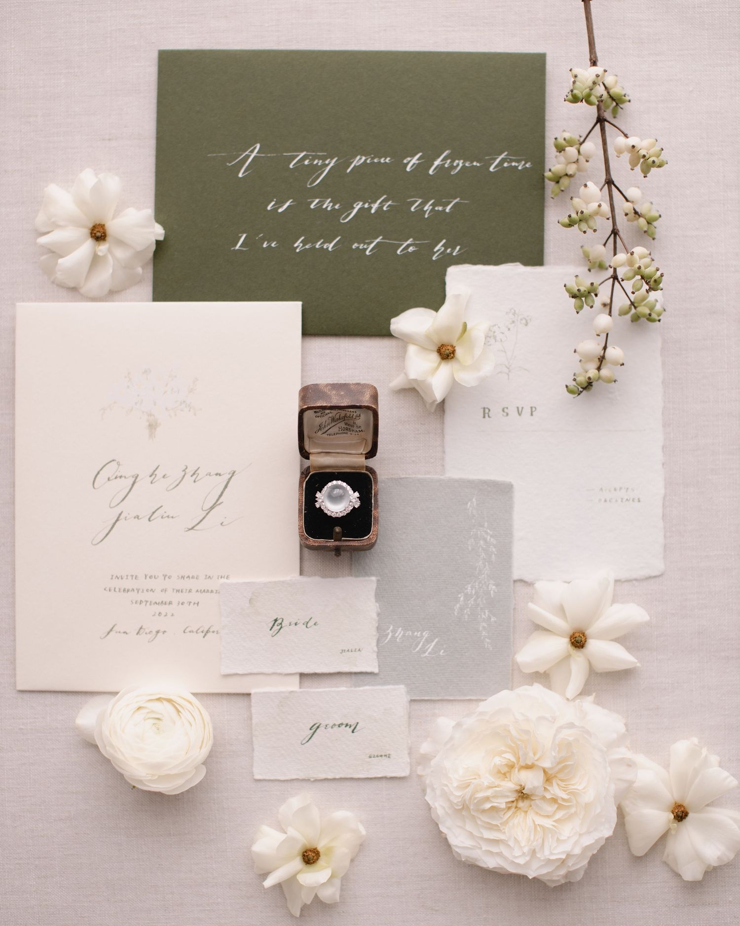 Wedding invitation suite with engagement ring, white flowers, and olive green accents.