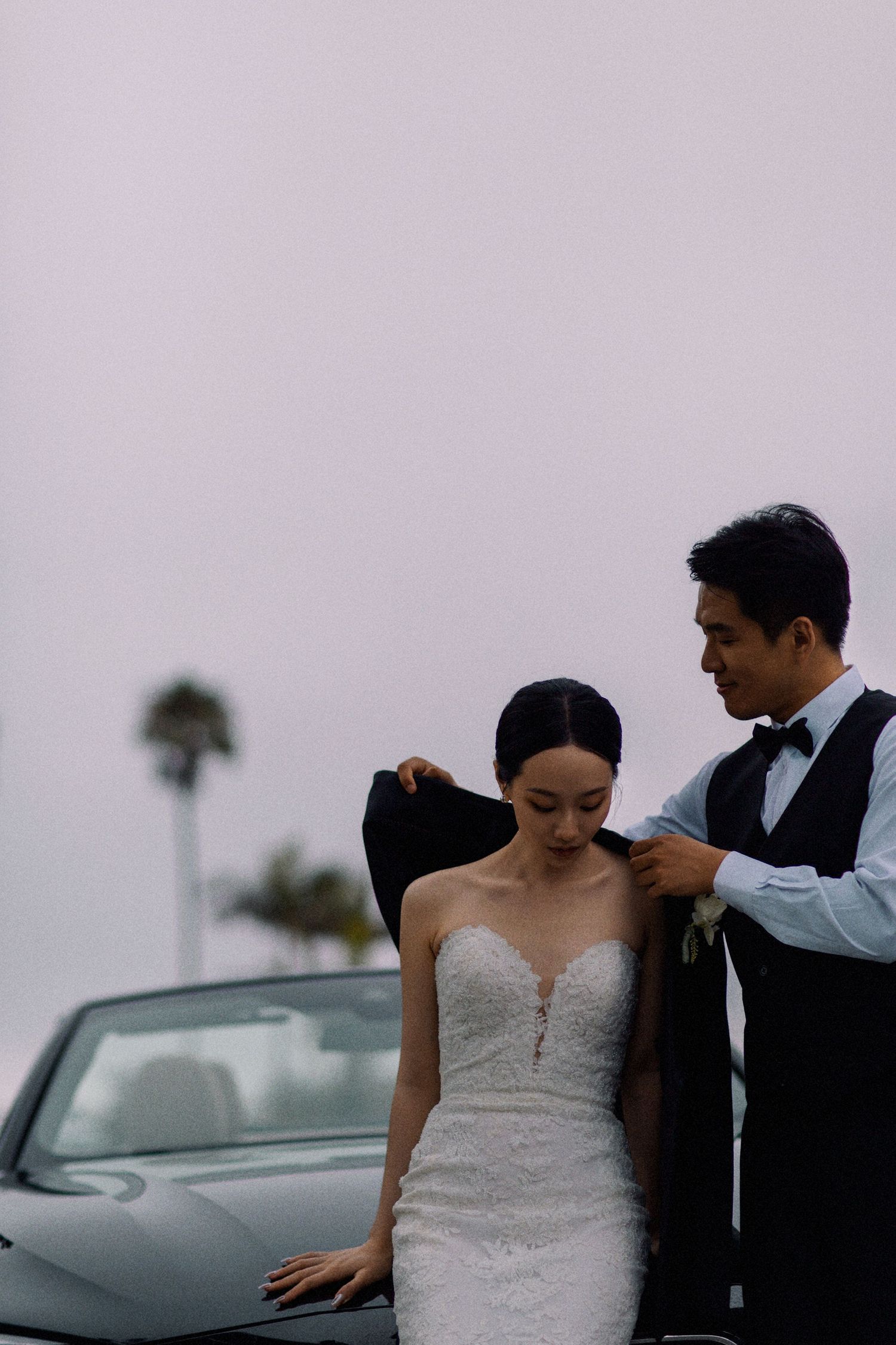 A groom helps his bride with her jacket next to a luxury car under an overcast sky.