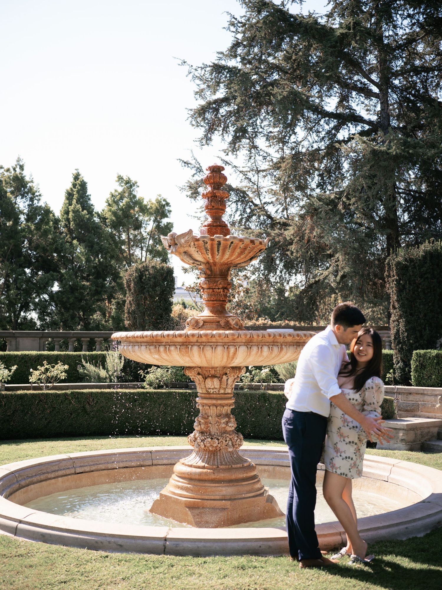 Ornate stone fountain with tiered basins stands as centerpiece in elegant garden with manicured hedges and cypress trees.